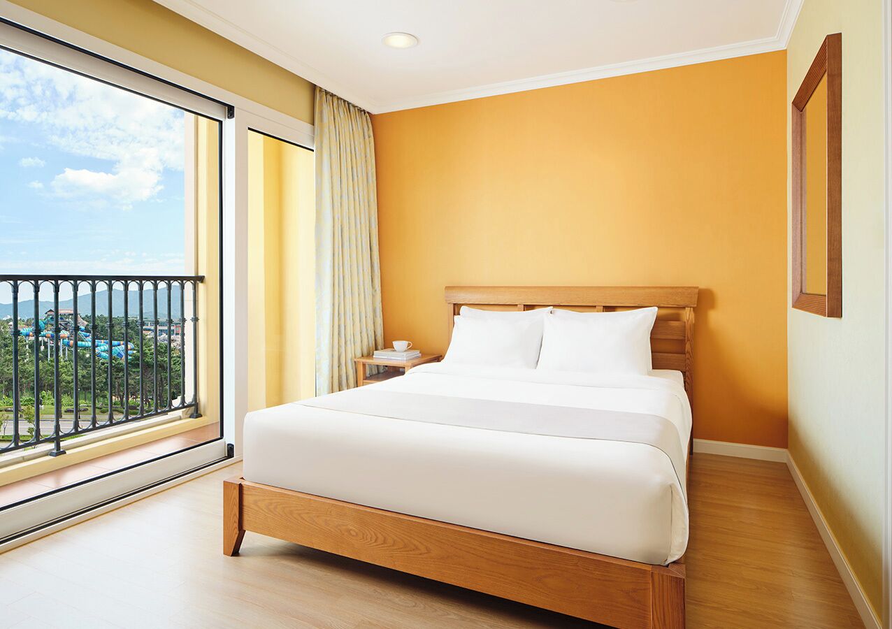 A hotel room with a large bed, a nightstand, a window with a view of a resort, and a balcony.