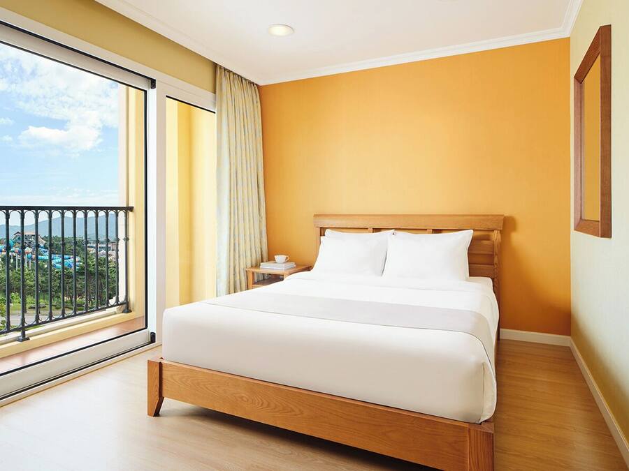 A hotel room with a large bed, a nightstand, a window with a view of a resort, and a balcony.