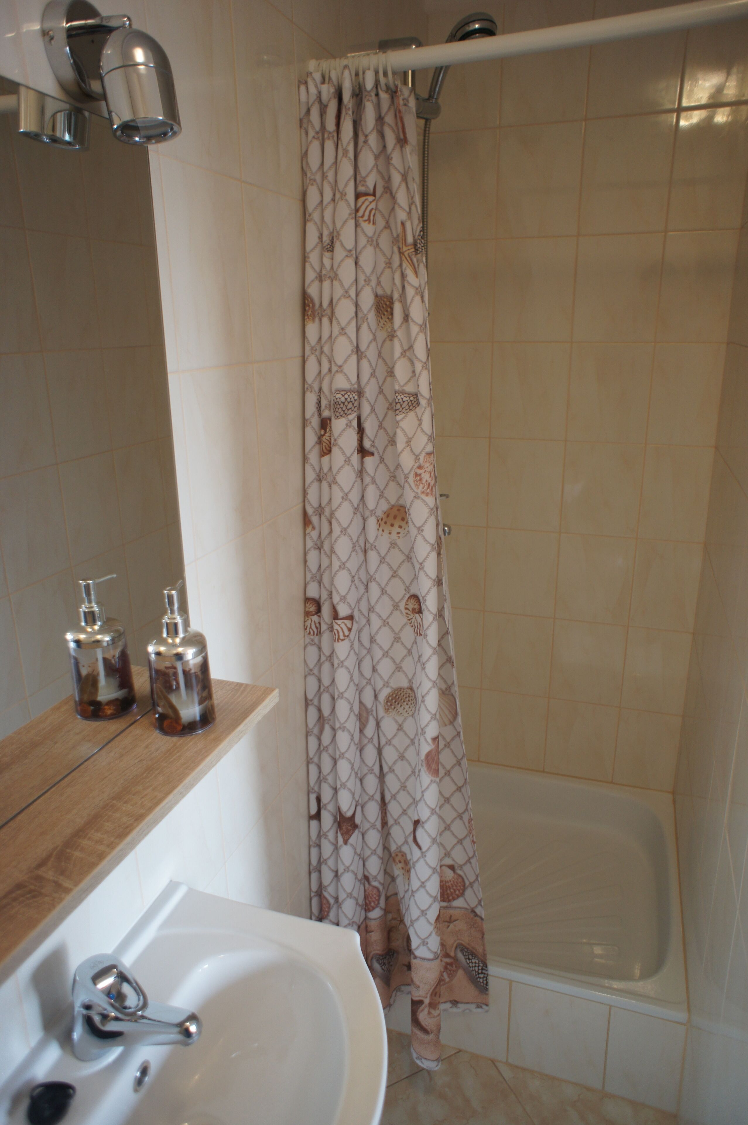Un baño con lavamanos, dos dispensadores de jabón, una cortina de ducha con estampado de conchas marinas y una bañera.