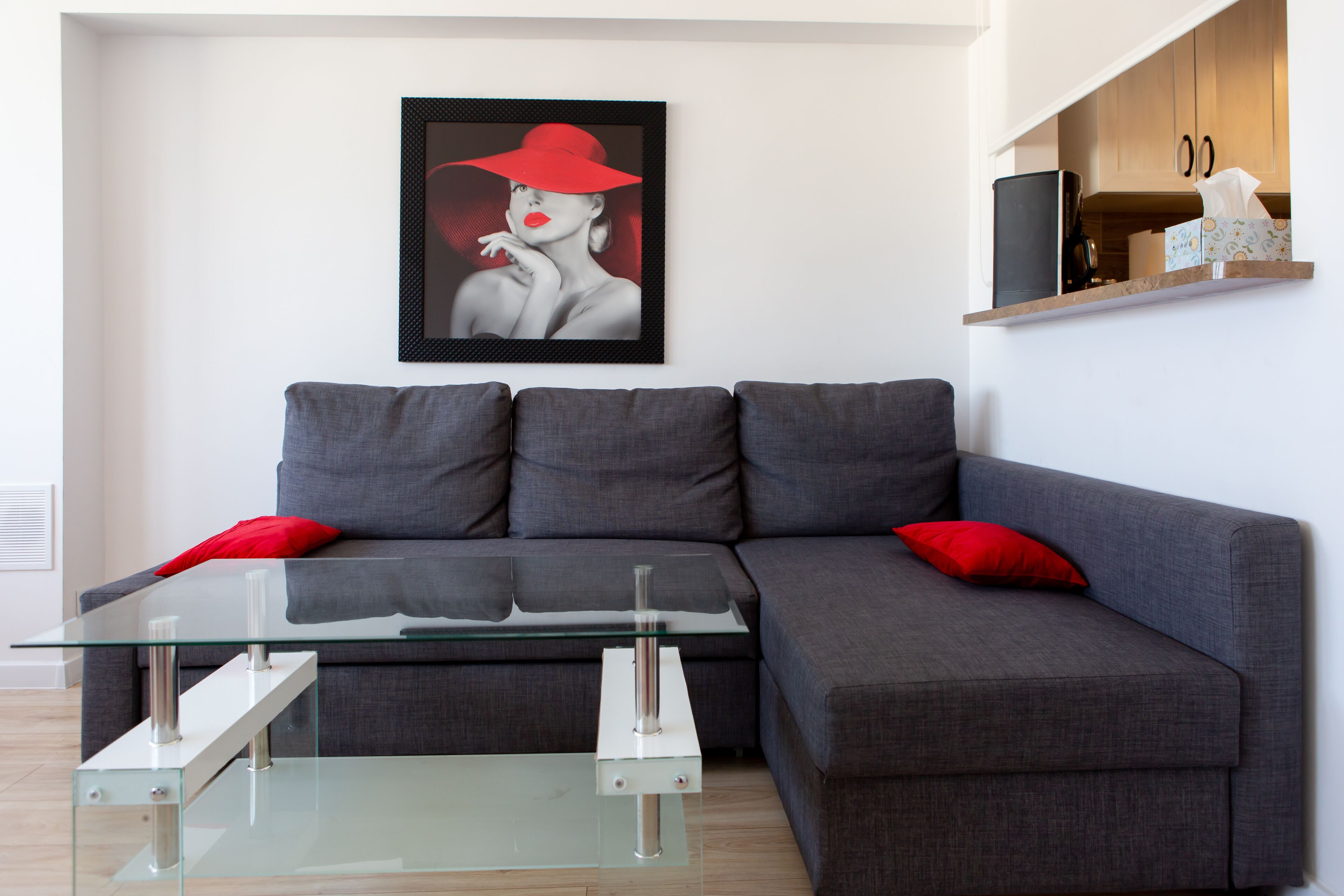Ein modernes Wohnzimmer mit grauem Sofa, roten Kissen, einem Glastisch und einem gerahmten Kunstwerk an der Wand.