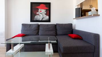 A modern living room with a grey sofa, red cushions, a glass coffee table, and a framed artwork on the wall.