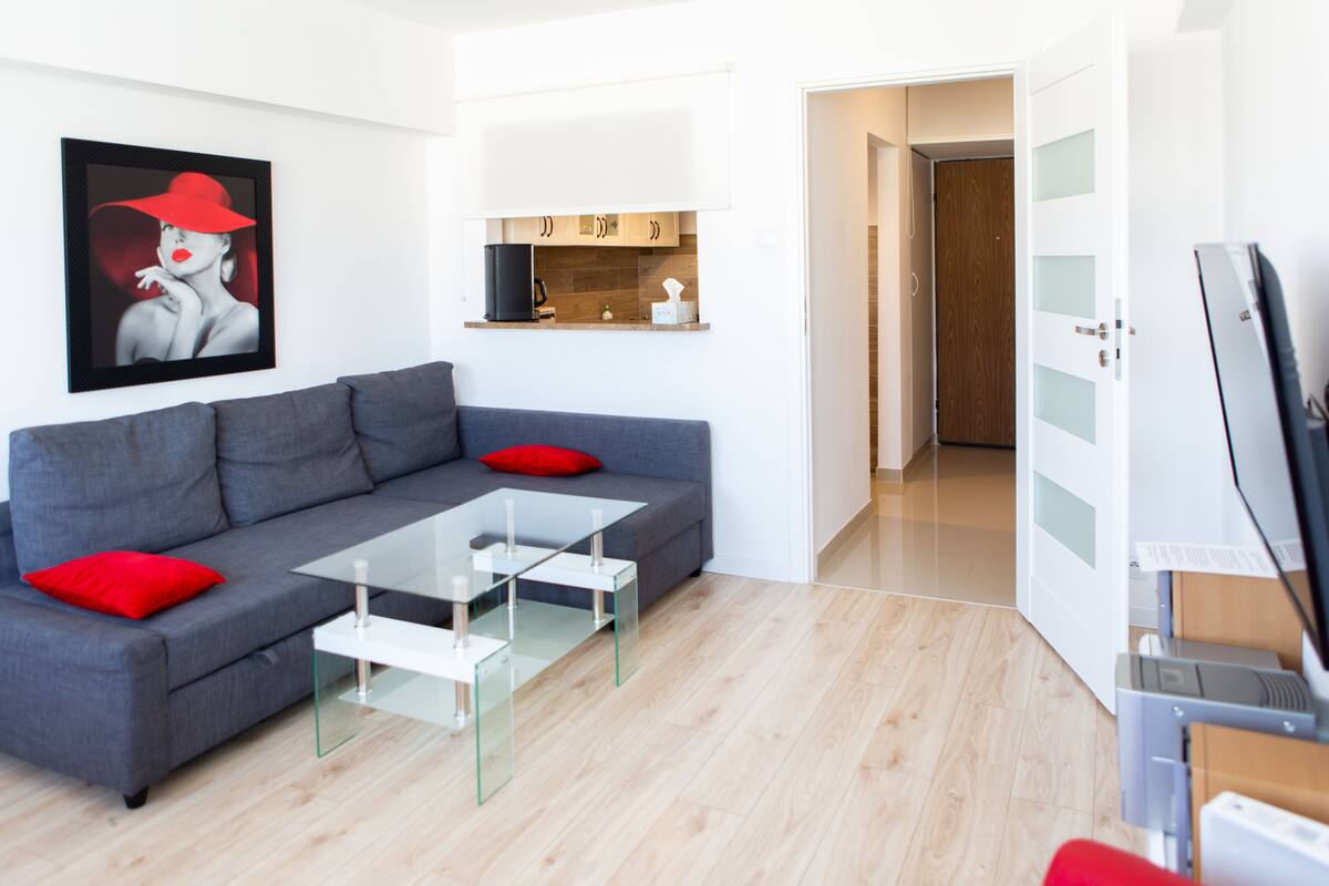 A modern living room with a grey sofa, red cushions, a glass coffee table, and a kitchen area with wooden cabinets and a white countertop.