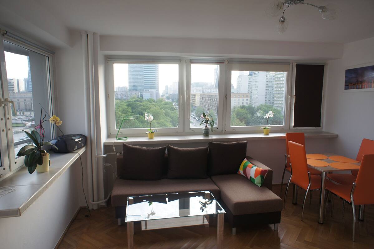 A modern living room with a sofa, coffee table, and dining area with chairs. Large windows offer a view of the cityscape.