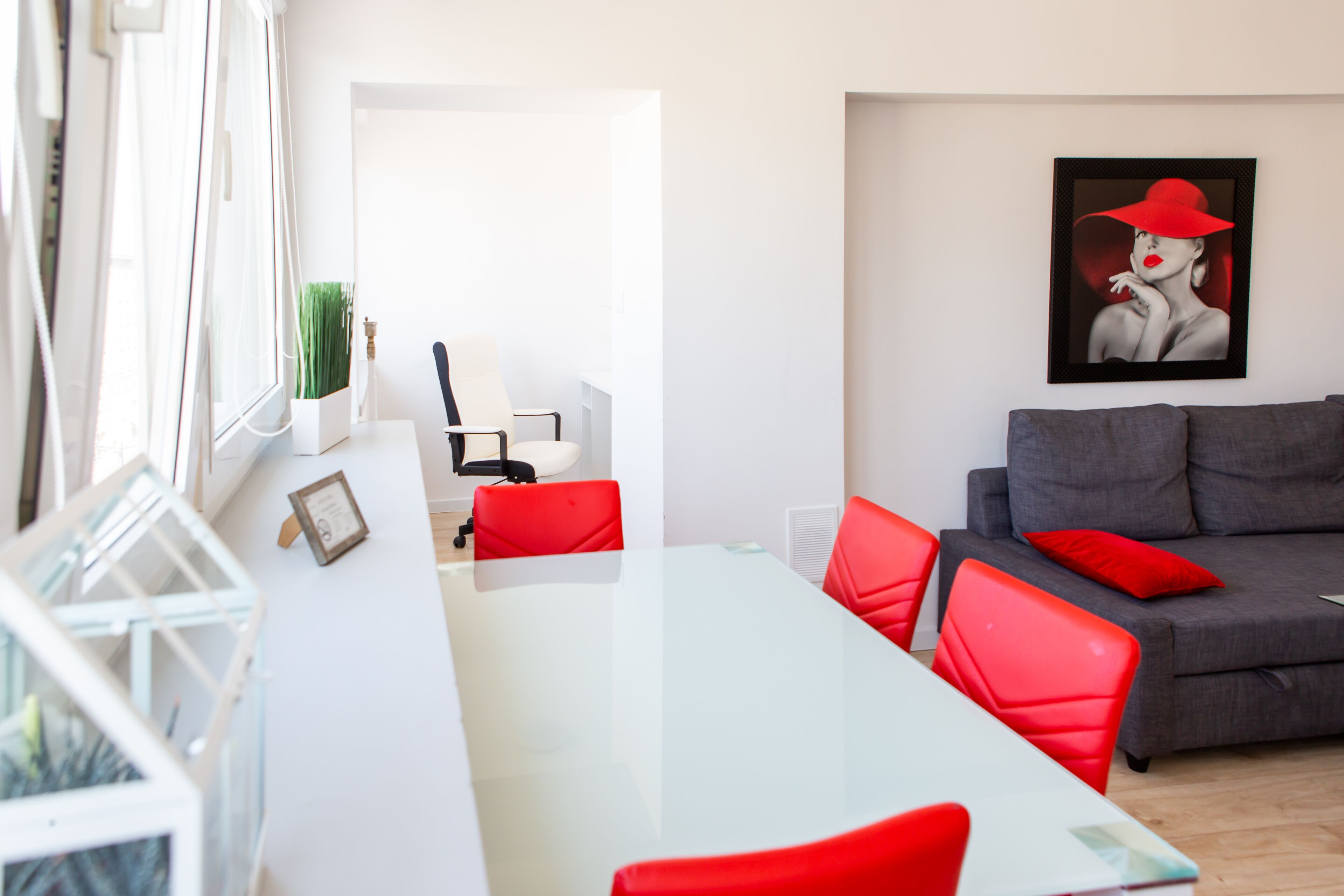 Ein modernes Wohnzimmer mit einem Glastisch, roten Stühlen, einem grauen Sofa mit roten Kissen und einem gerahmten Kunstwerk an der Wand.