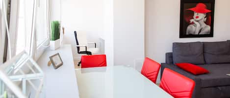 Ein modernes Wohnzimmer mit einem Glastisch, roten Stühlen, einem grauen Sofa mit roten Kissen und einem gerahmten Kunstwerk an der Wand.