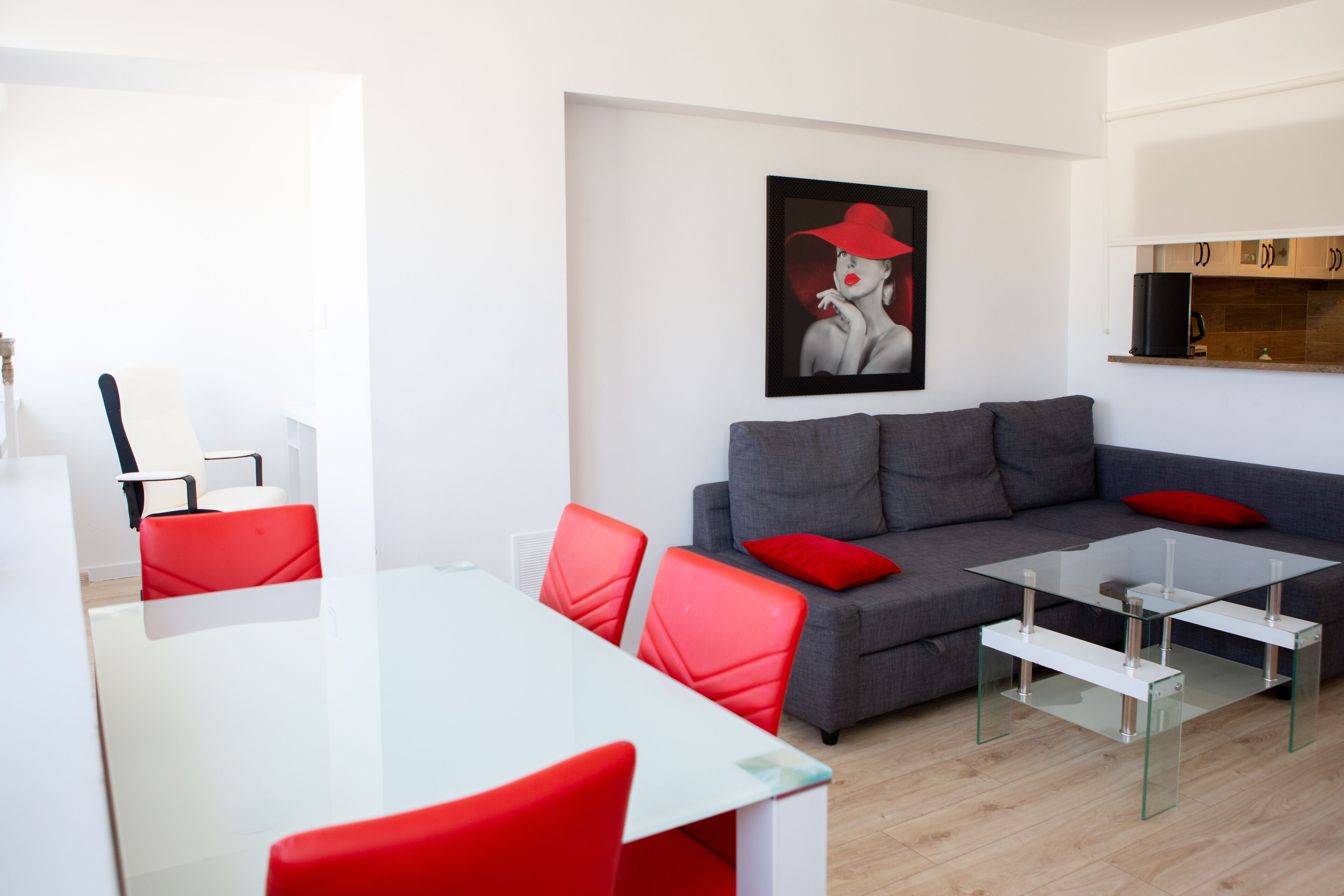 Ein modernes Wohnzimmer mit einem Glastisch, roten Stühlen, einem grauen Sofa mit roten Kissen und einem gerahmten Kunstwerk an der Wand.