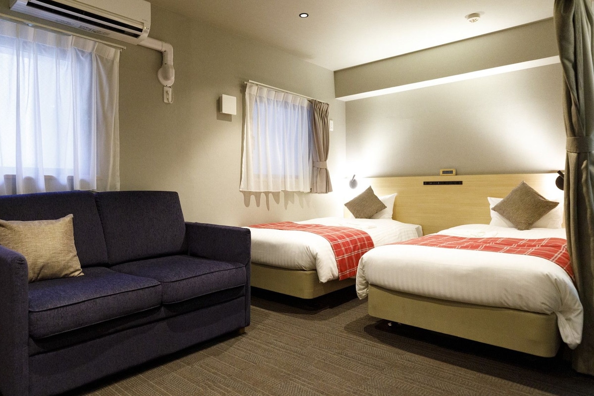 Quarto de hotel com sofá, duas camas e cortinas.