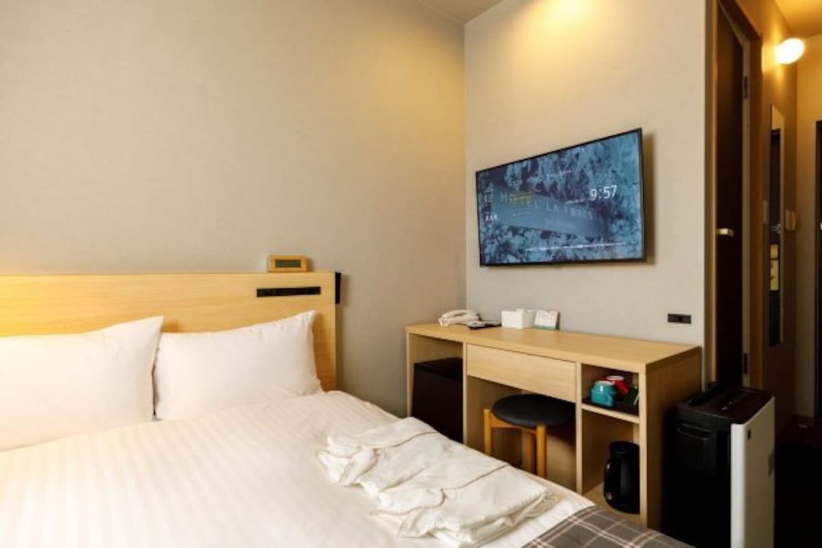 A hotel room with a bed, a desk, a chair, a coffee machine, and a television displaying a snowy landscape.