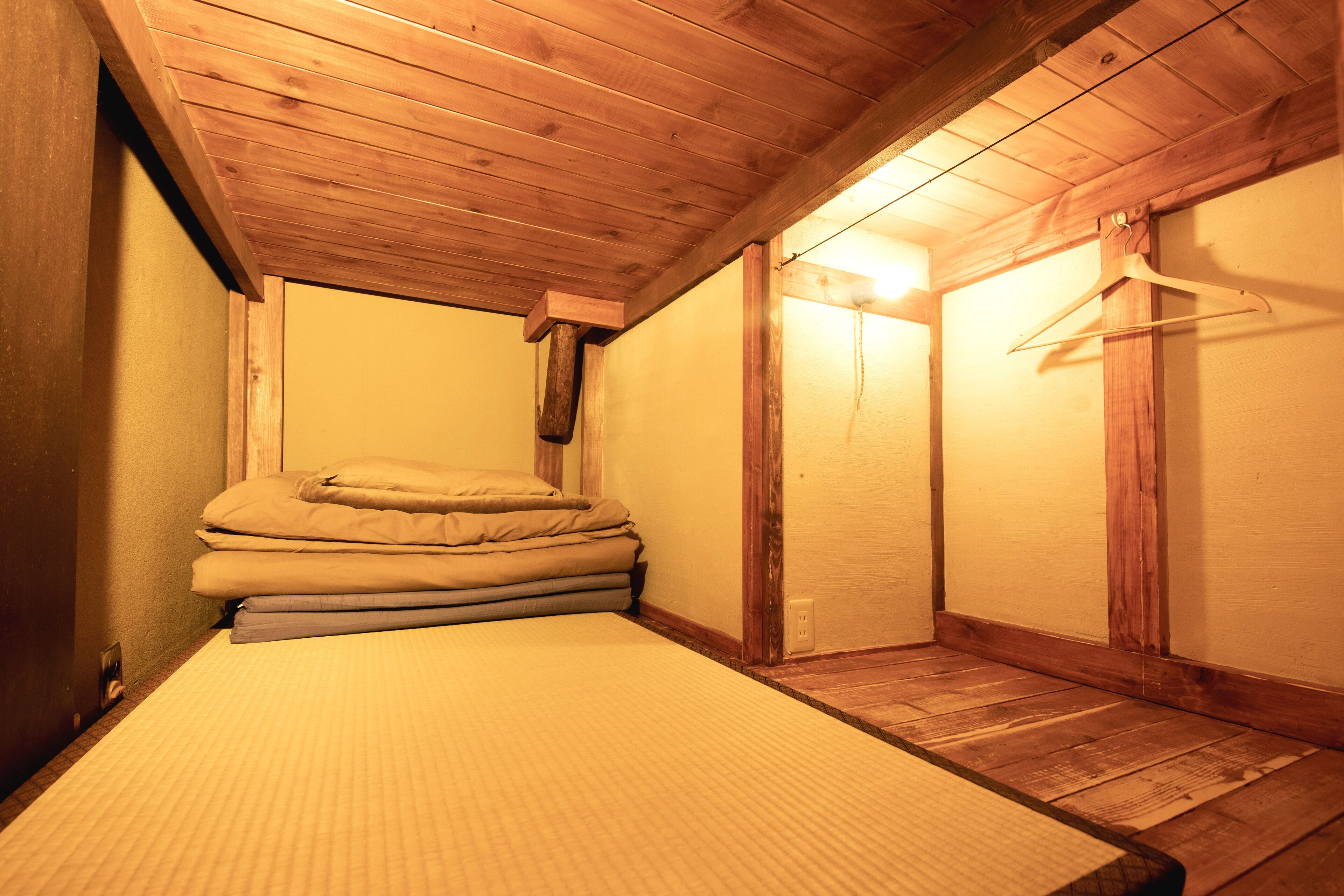 A small, wooden room with a bed, a hanging coat rack, and a wall-mounted light fixture.