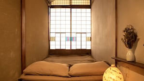 A traditional Japanese room with tatami flooring, a low bed with two pillows, a shoji screen window, and a vase with dried flowers on a shelf. - Guesthouse YULULU (Kyoto)