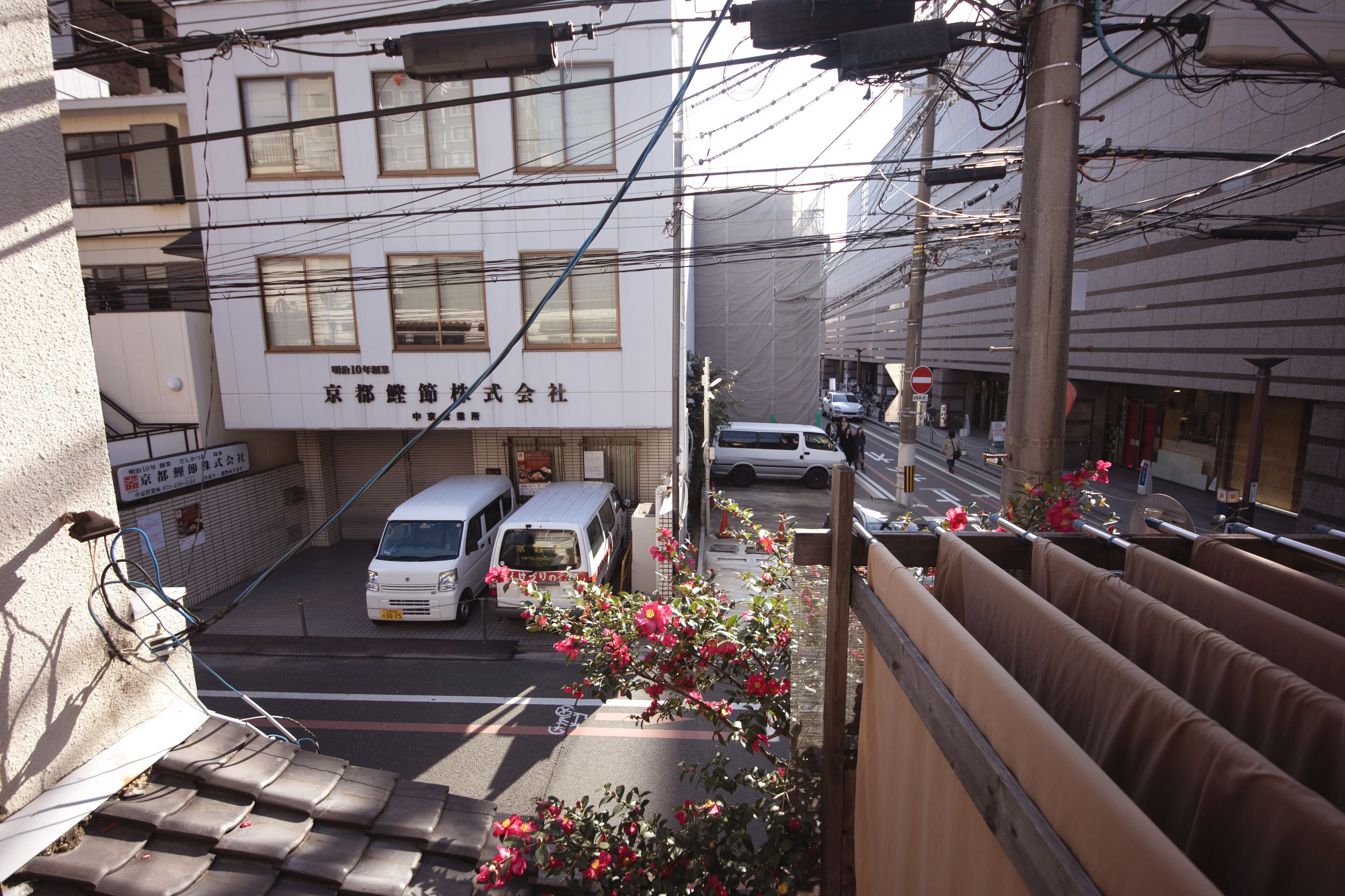 A building with multiple floors, a white facade, and a sign with Japanese characters. There are parked vehicles in front, and a street with a pedestrian crossing.