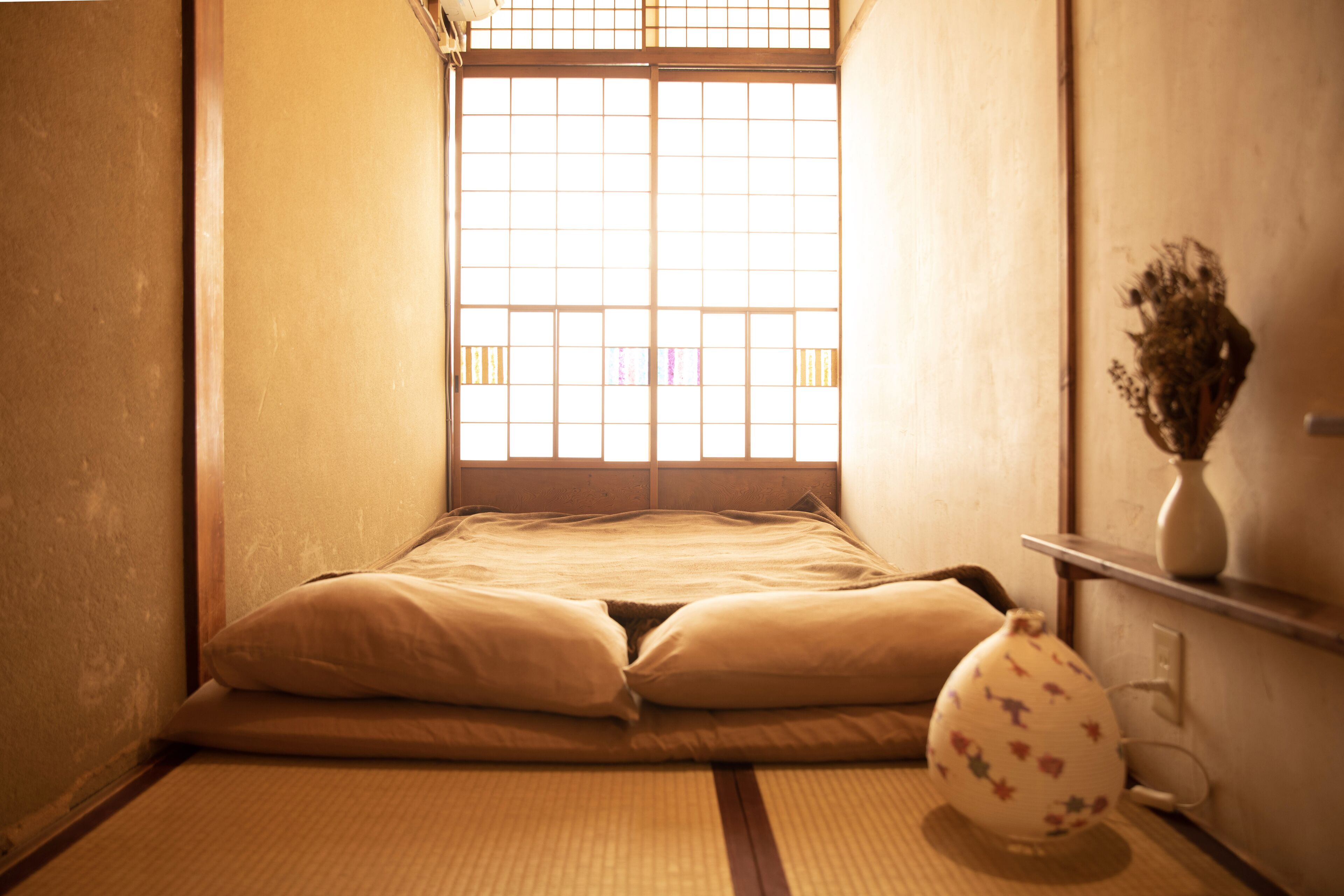 A tatami mat room with a futon bed, a vase with dried flowers, and a sliding door with a grid pattern.