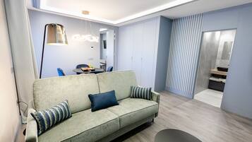 A modern living room with a green sofa, a dining table with chairs, and a kitchen area in the background.