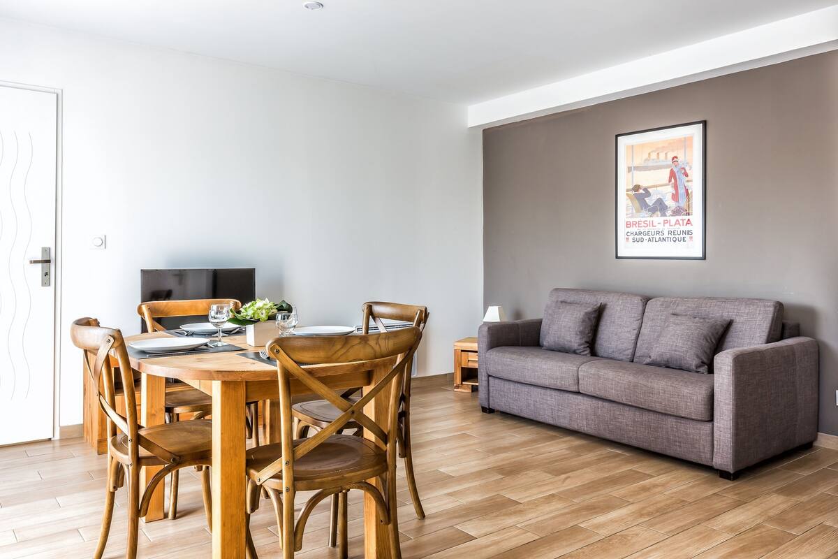 A modern living room with a grey sofa, wooden dining table with chairs, and a television on a stand.