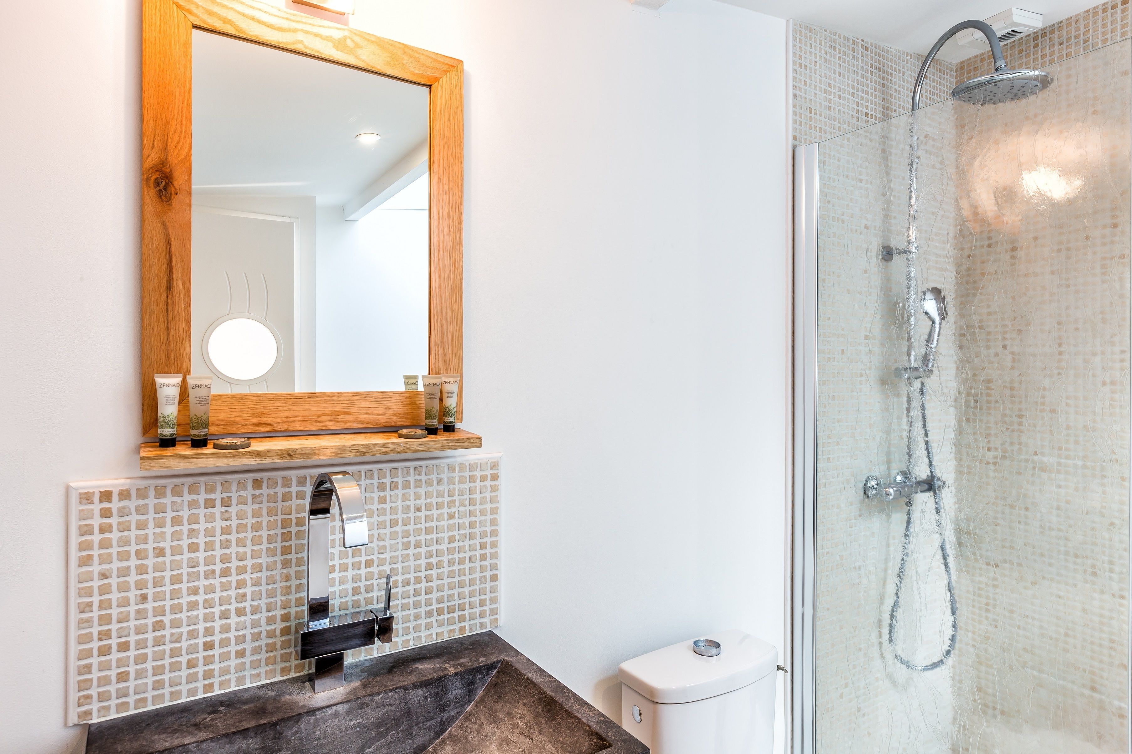 A bathroom with a wooden-framed mirror, a tiled backsplash, a sink with a dark countertop, and a glass-enclosed shower with a handheld showerhead.