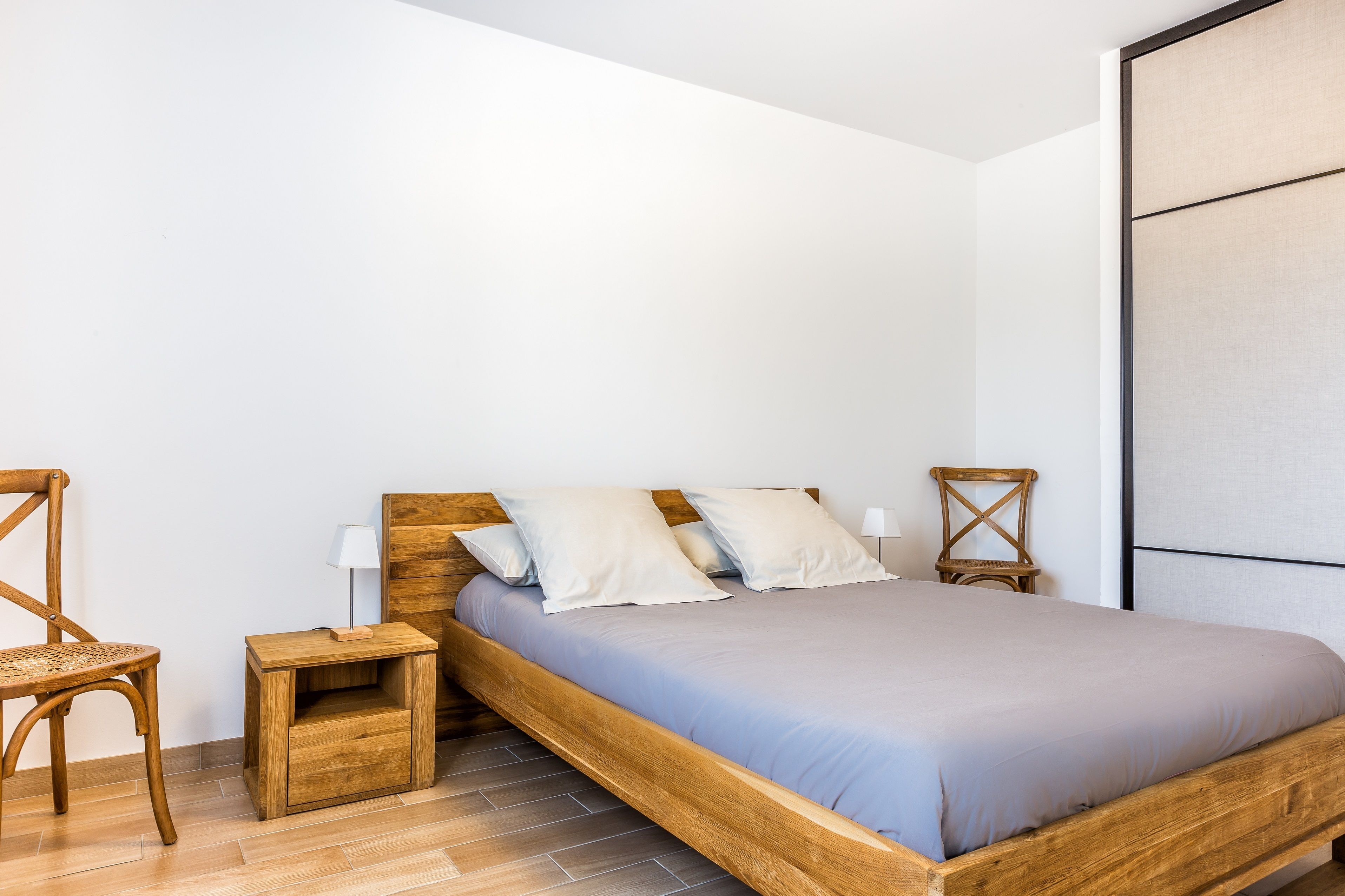 A bedroom with a wooden bed, two pillows, a nightstand with a lamp, and a chair.