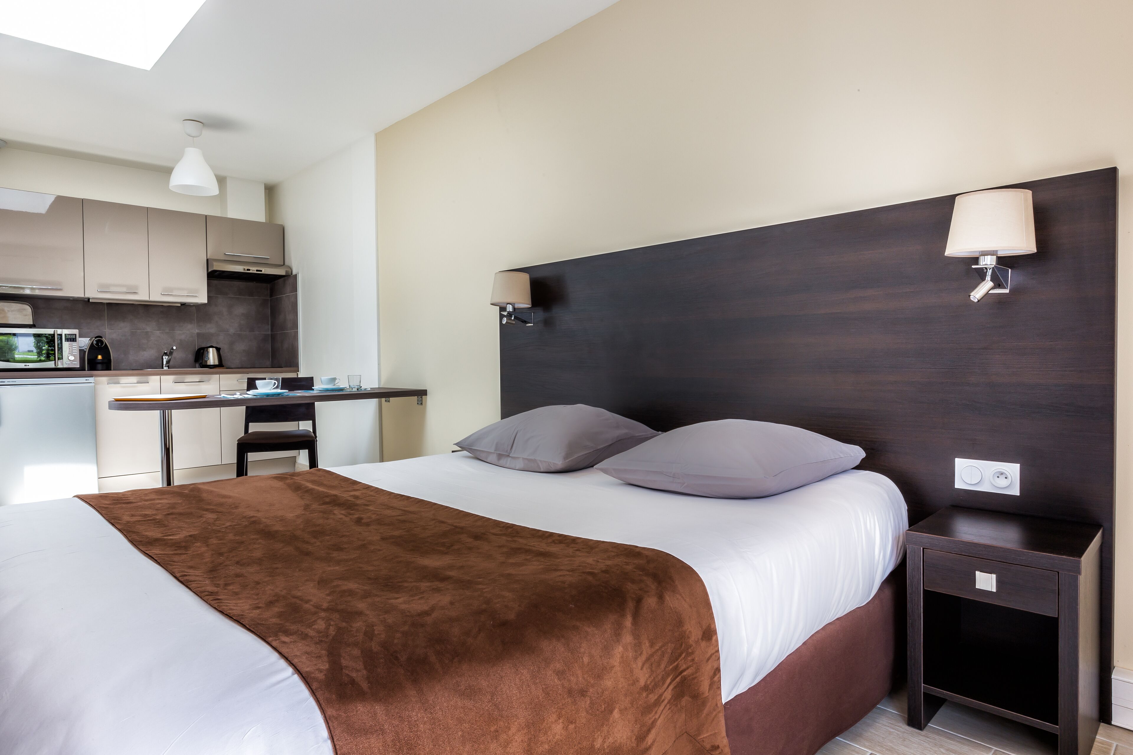 A hotel room with a bed, bedside table, and a view of the kitchen.