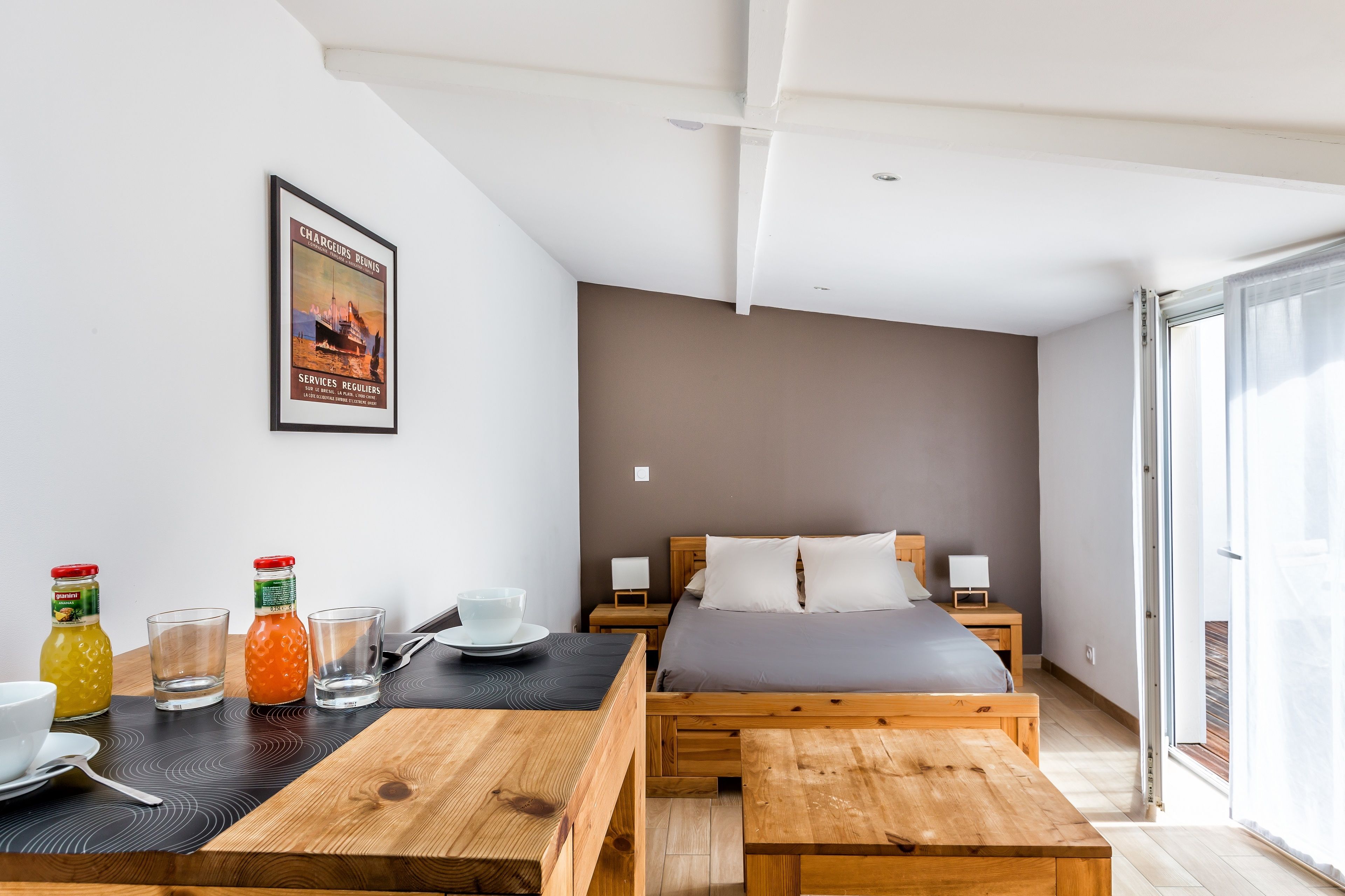 A modern bedroom with a bed, bedside tables, a wooden table with drinks, and a framed picture on the wall.