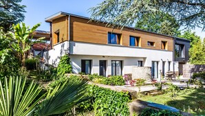A modern two-story house with a wooden facade, large windows, and a well-maintained garden with various plants and a patio area.