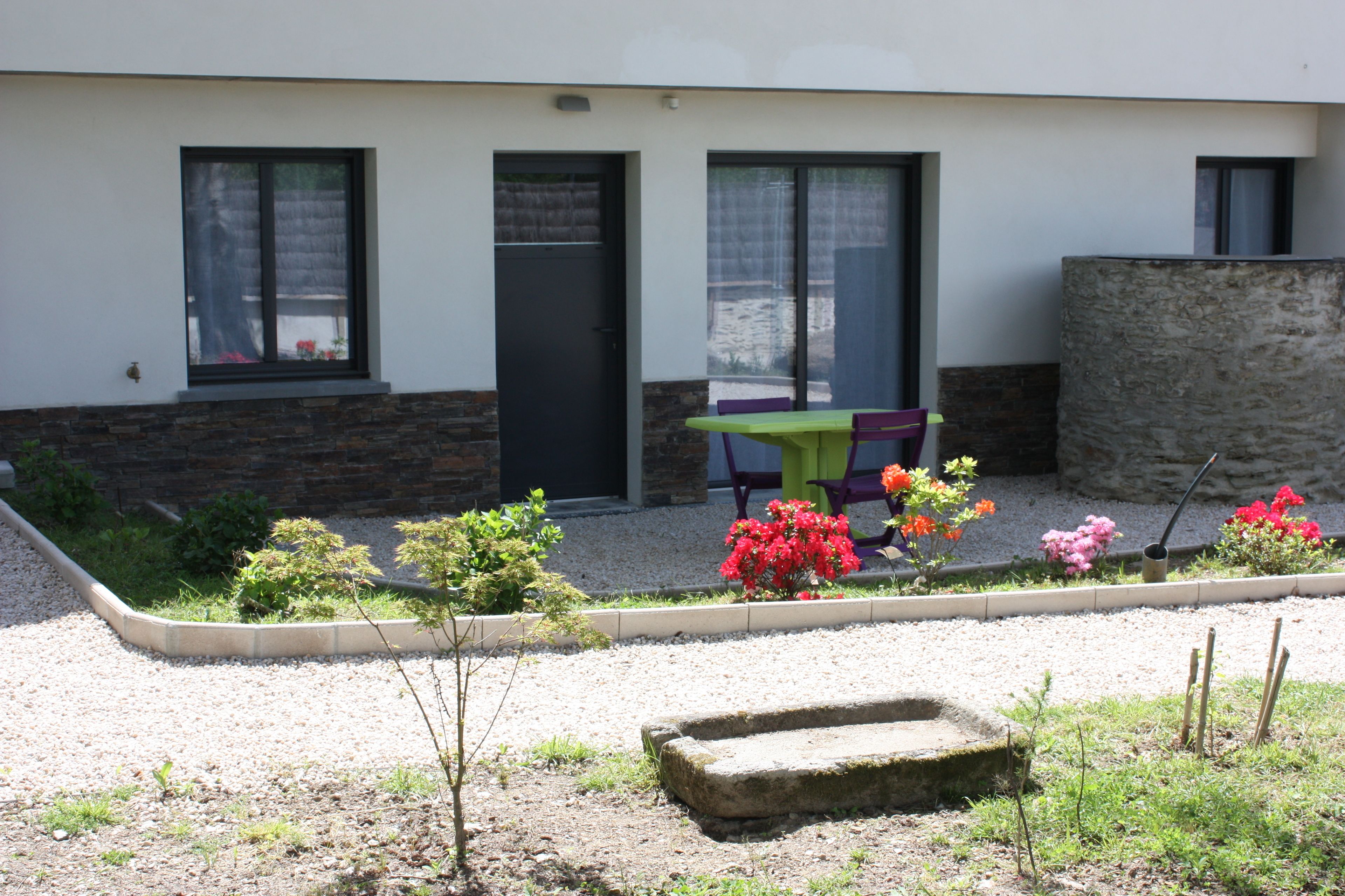 A modern building with a gravel courtyard, a small garden with colorful flowers, and a stone bench.
