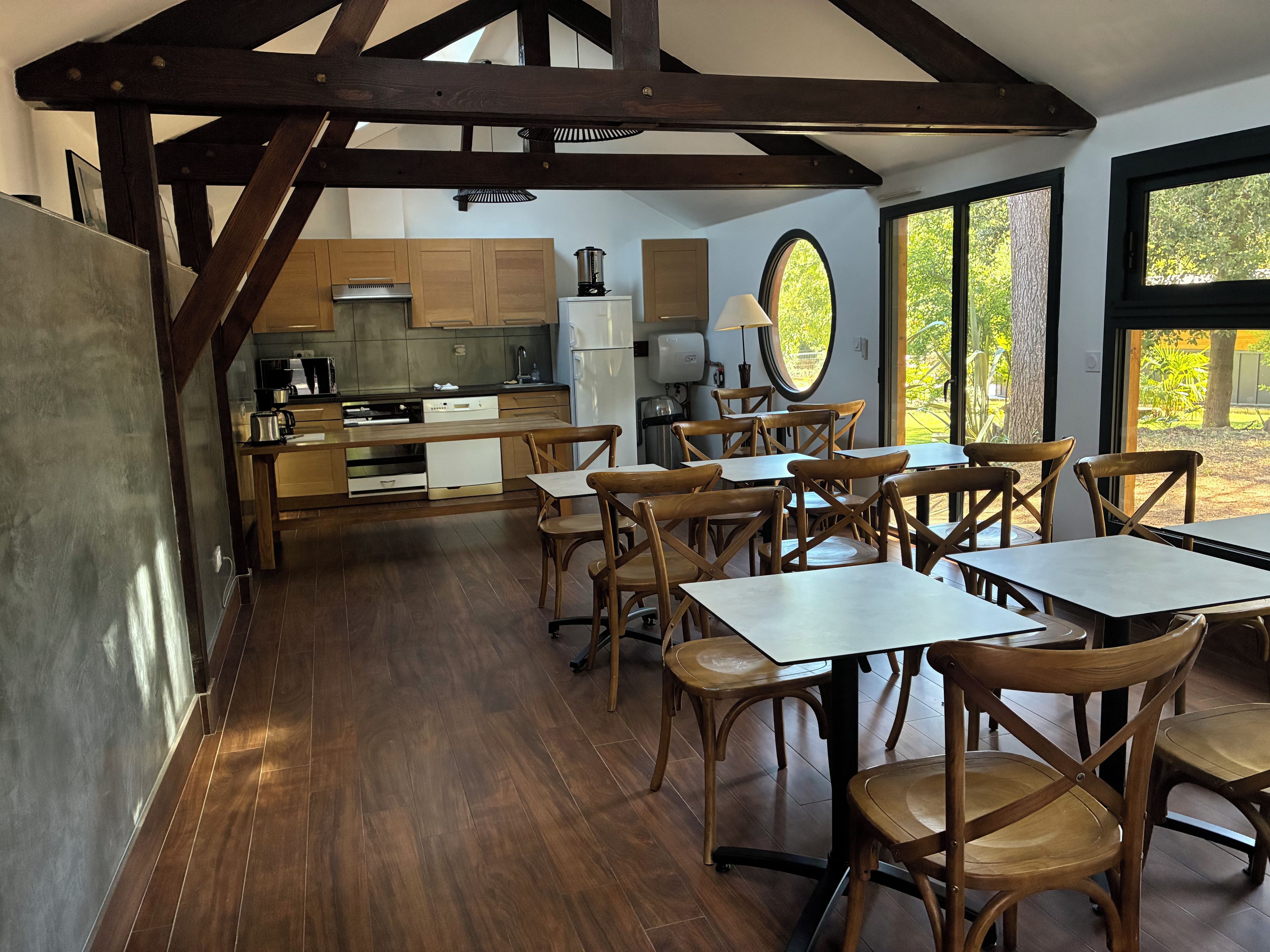 A dining area with wooden tables and chairs, a kitchen in the background, and large windows with a view of greenery.