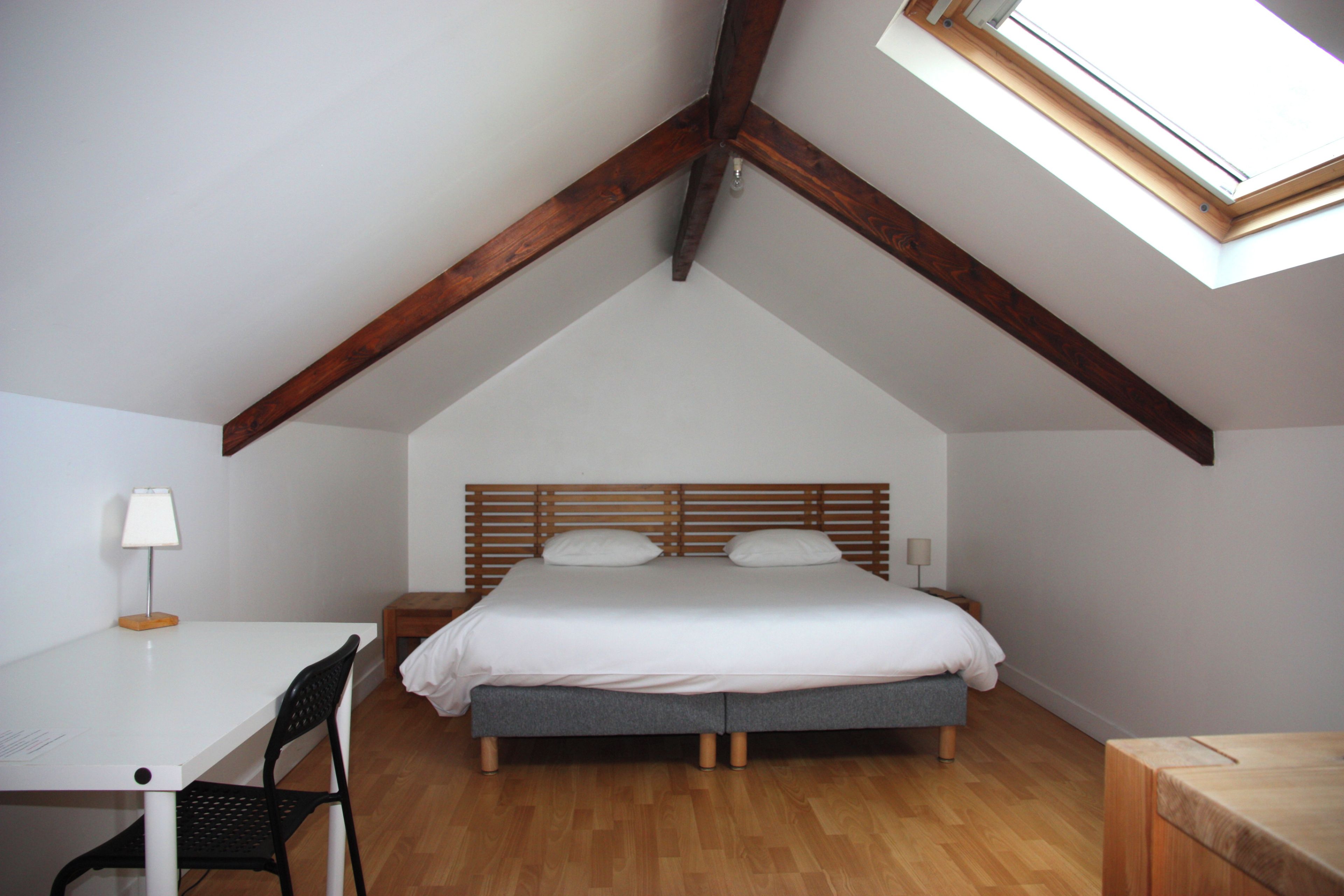 A bedroom with a sloped ceiling, a bed with a wooden headboard, a desk with a chair, and a small table.