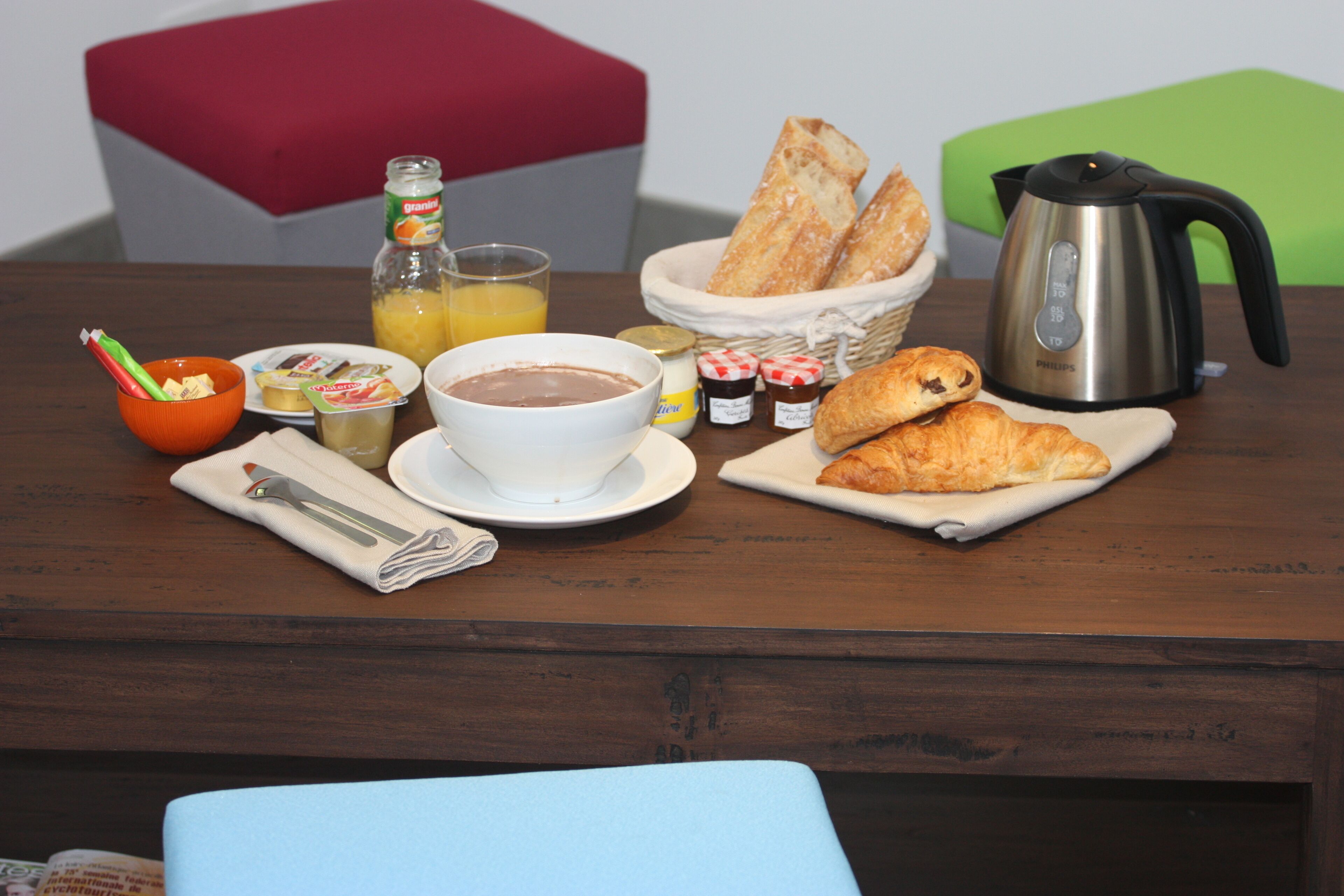 A breakfast table with bread, croissants, a coffee pot, a bottle of orange juice, and a bowl of cereal.
