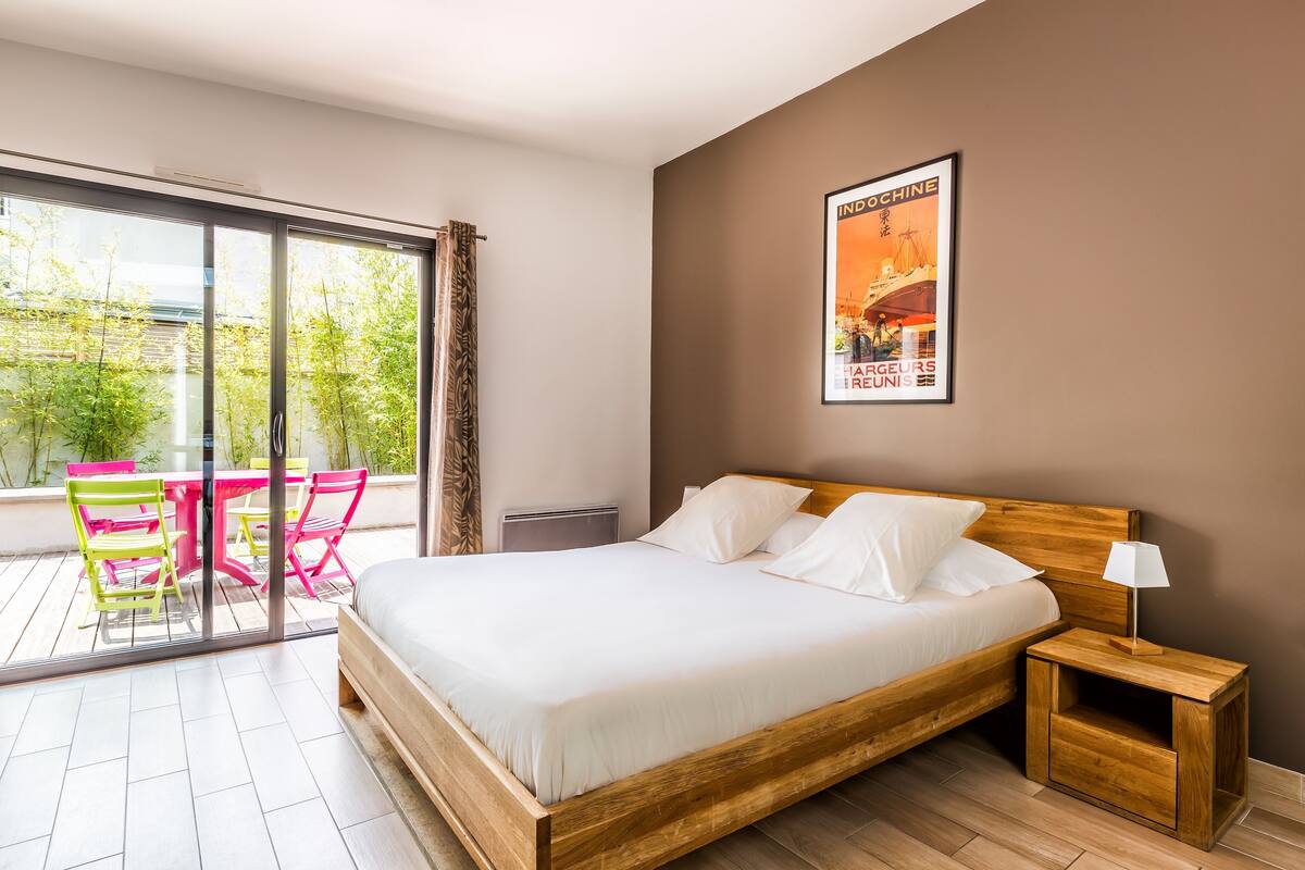 A bedroom with a wooden bed, a nightstand with a lamp, a window with curtains, and a poster on the wall.