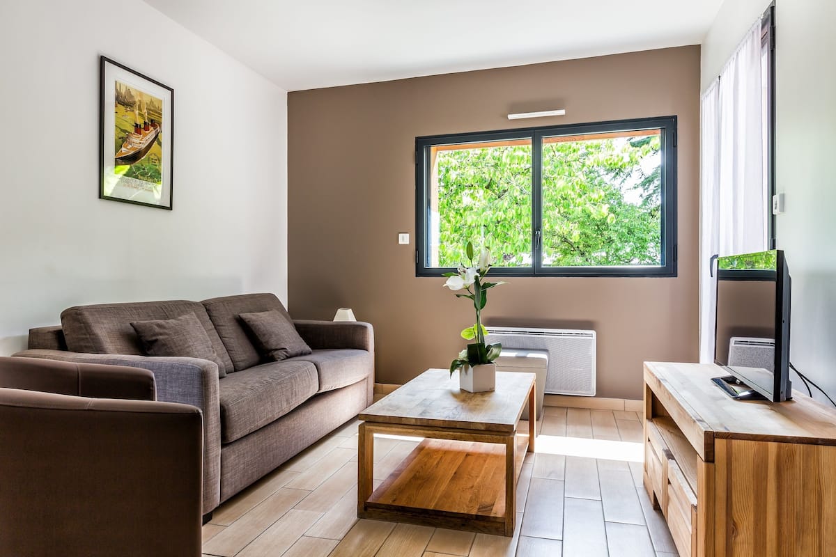 A modern living room with a sofa, coffee table, and TV stand, featuring a large window with a view of trees.