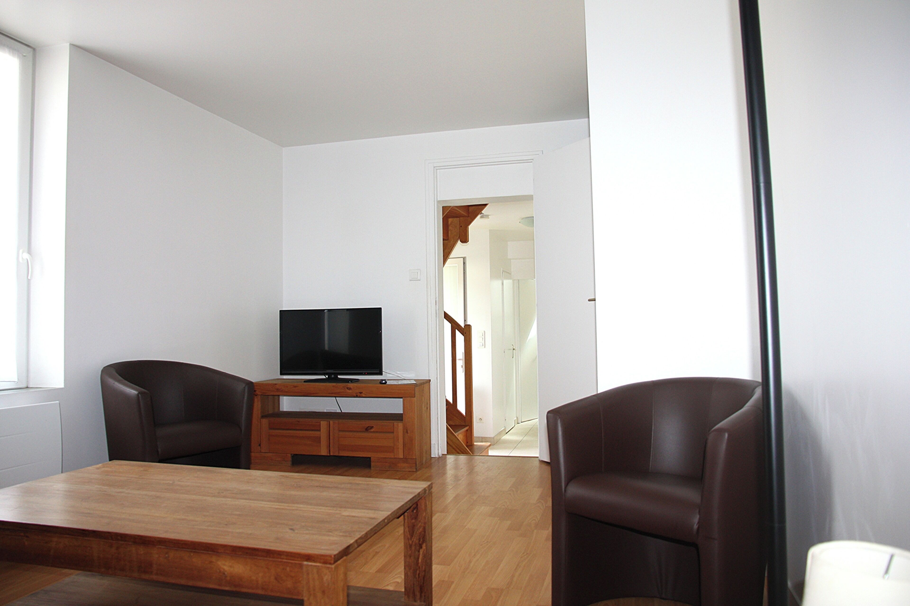 A modern living room with a wooden coffee table, two brown leather chairs, a flat-screen TV on a wooden stand, and a staircase leading to another room.