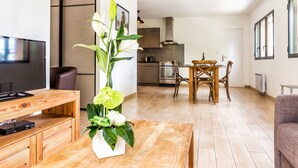 A modern living room with a wooden coffee table, a TV, and a floral centerpiece. A dining area with a wooden table and chairs is visible in the background.