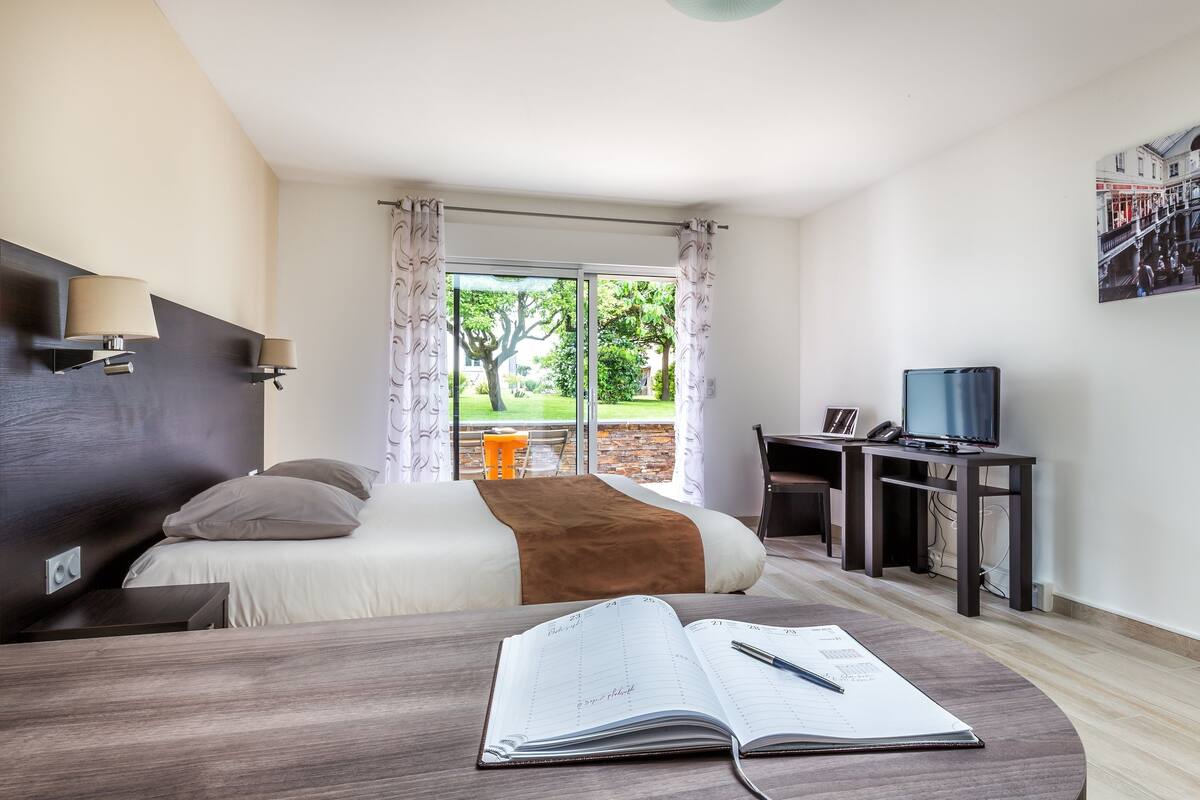 A hotel room with a bed, a desk with a TV, a window with curtains, and a calendar on the desk.