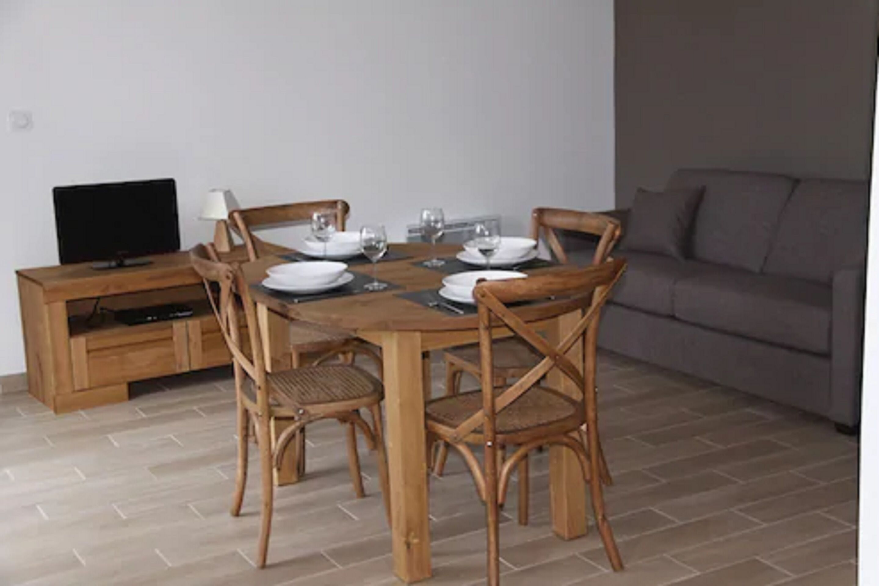 A dining area with a wooden table set for four, surrounded by wicker chairs. A television is on a wooden cabinet to the left.