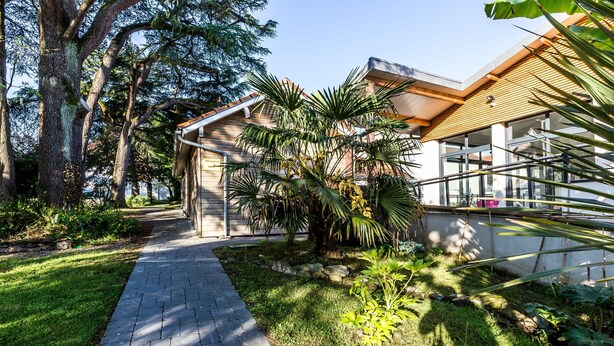 A modern building with a paved pathway leading to a covered entrance, surrounded by lush greenery and large trees.