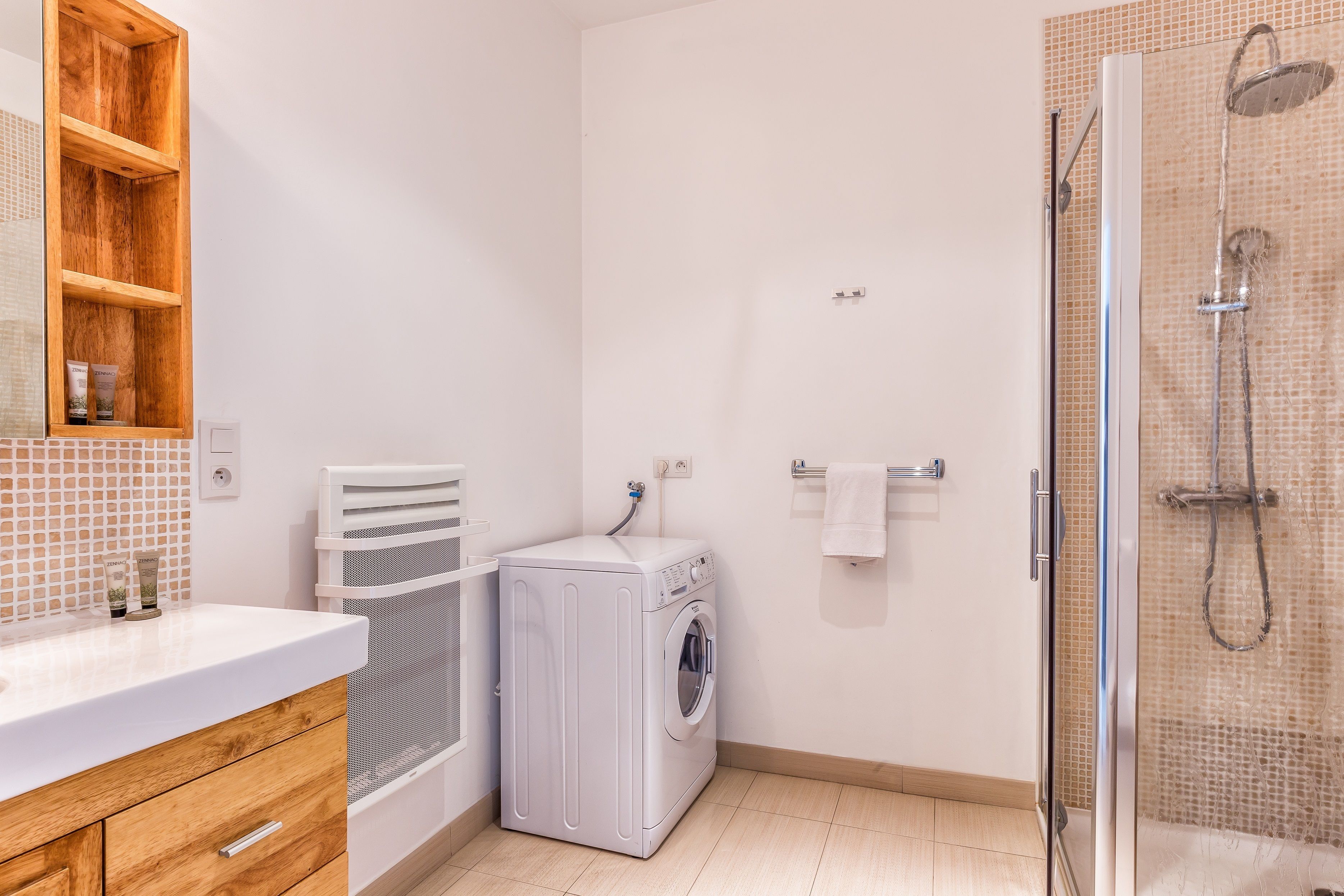 A compact bathroom with a washing machine, a glass shower enclosure, and a wooden cabinet with toiletries.