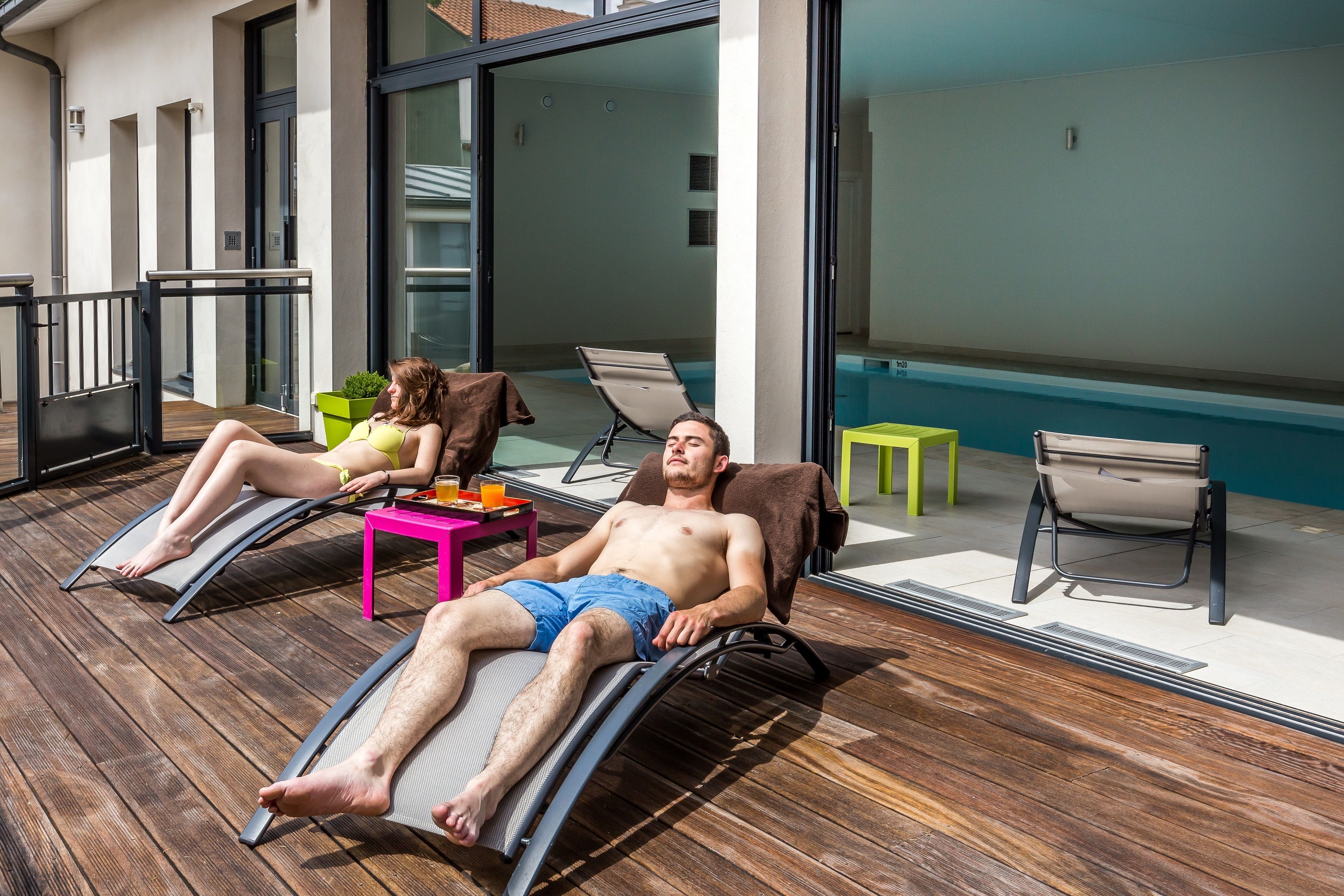 A sunlit outdoor area with two lounge chairs, a small table, and a pool in the background.