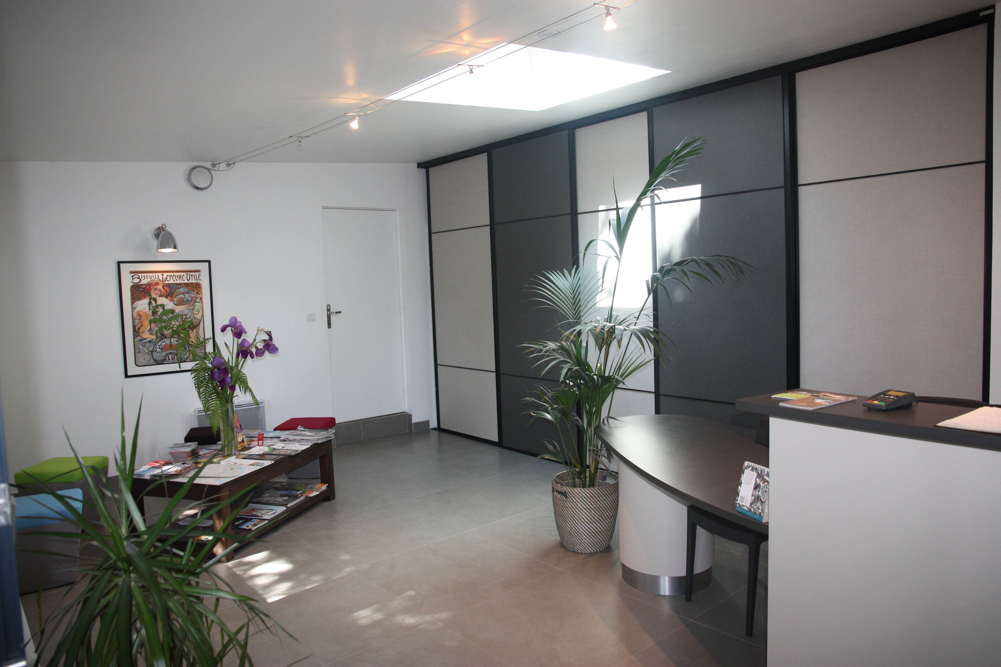 Un espace d’accueil moderne comprenant un comptoir de réception, une grande plante en pot, une œuvre d’art encadrée au mur et un coin salon avec une table et des chaises.