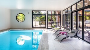 An indoor swimming pool with clear glass walls, a tiled floor, and lounge chairs.