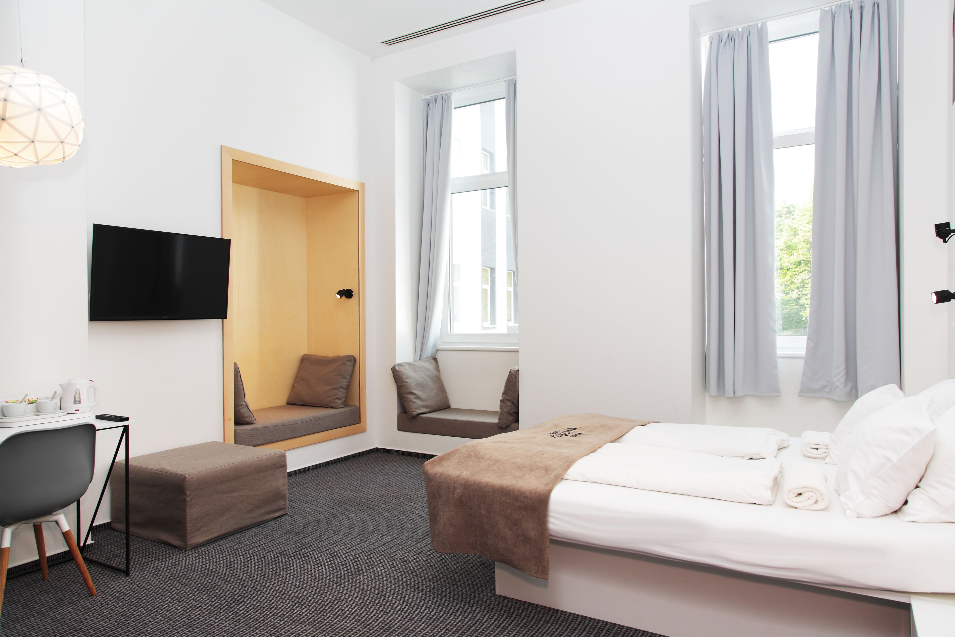 A modern hotel room with a bed, a desk with a chair, a sofa, and a TV mounted on the wall.