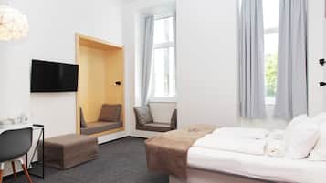 A modern hotel room with a bed, a desk with a chair, a sofa, and a TV mounted on the wall.