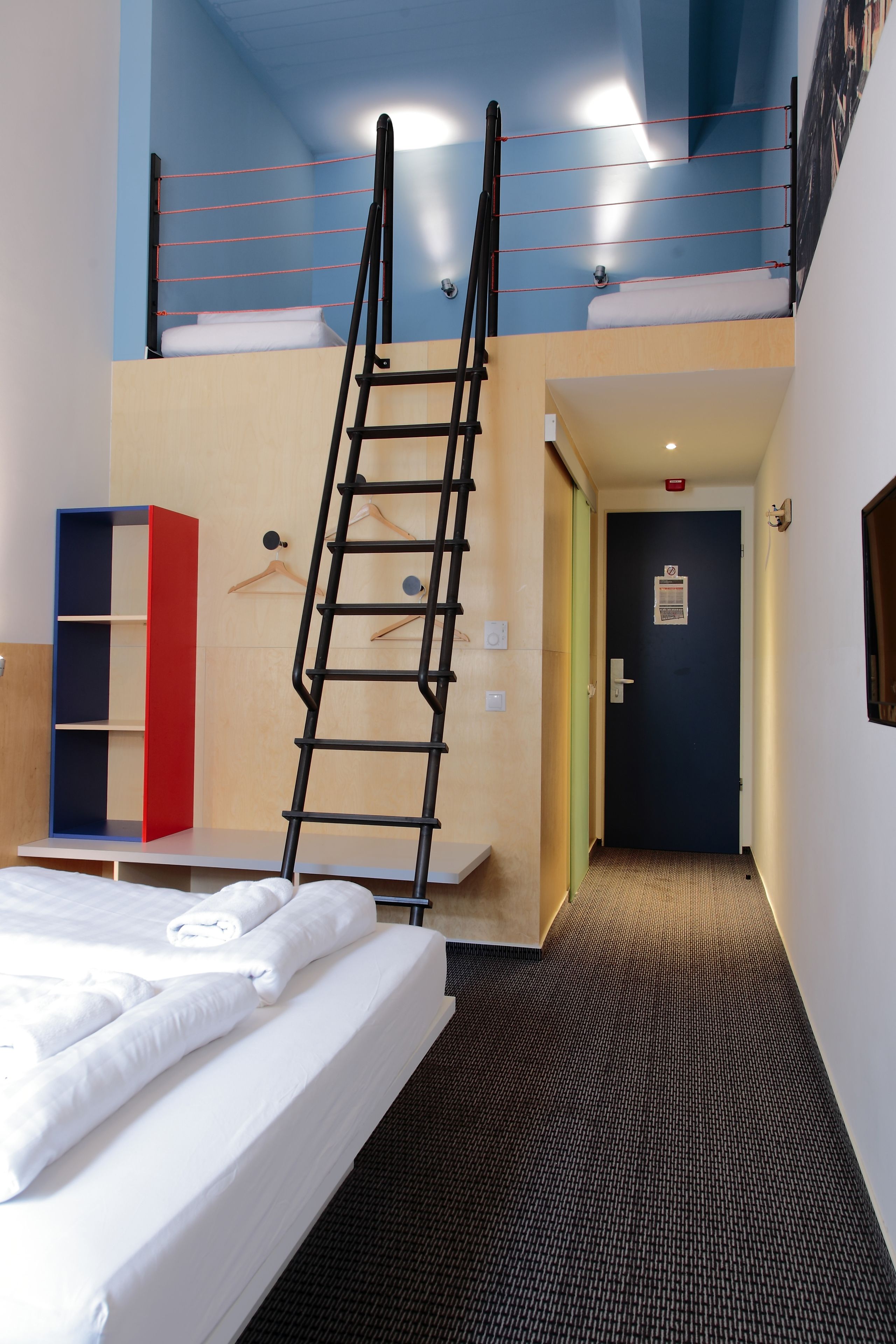 A hotel room with a bunk bed, a ladder, a desk, and a wardrobe.