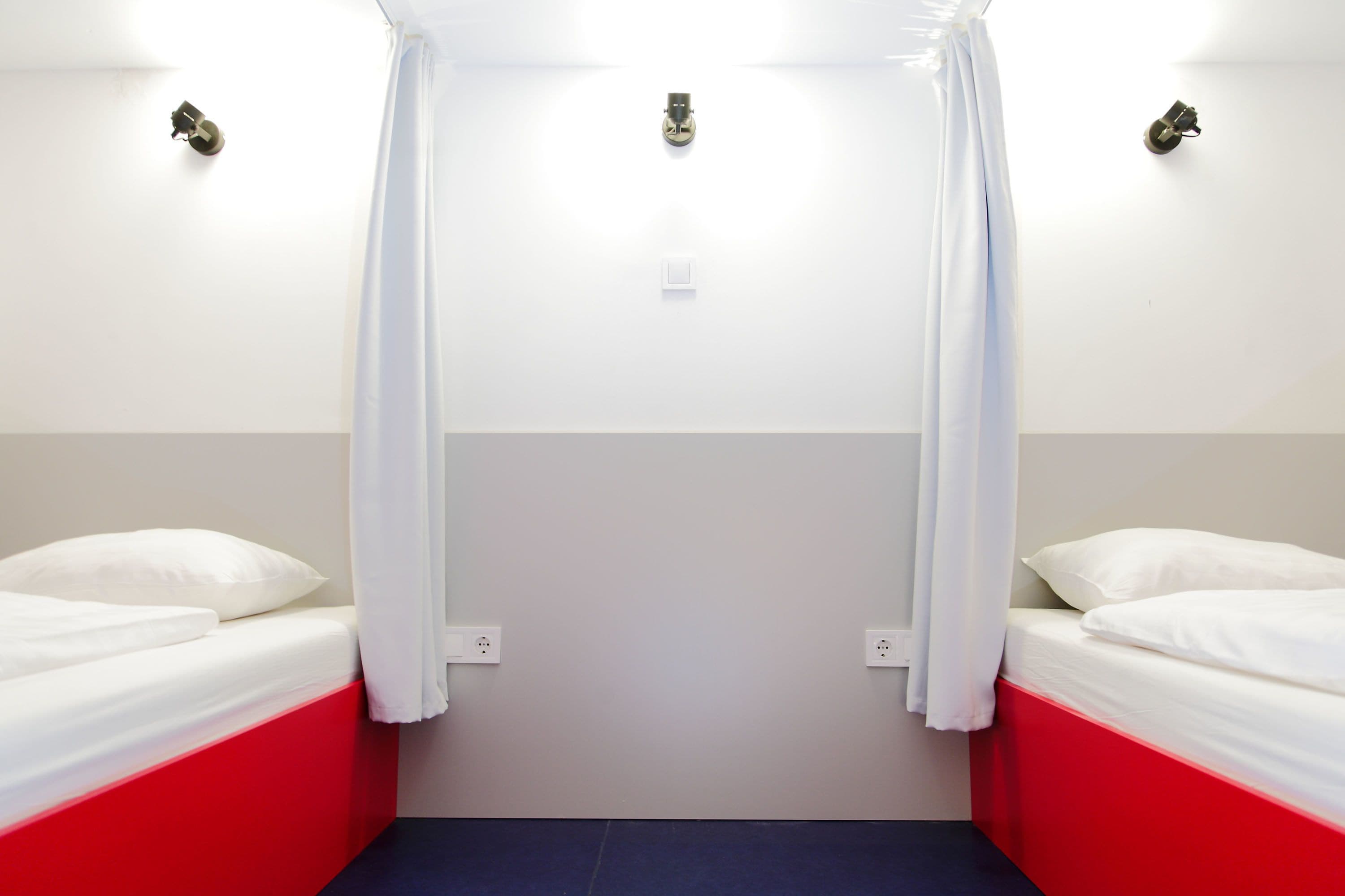A room with two beds, each with white bedding and red headboards, separated by a white curtain.