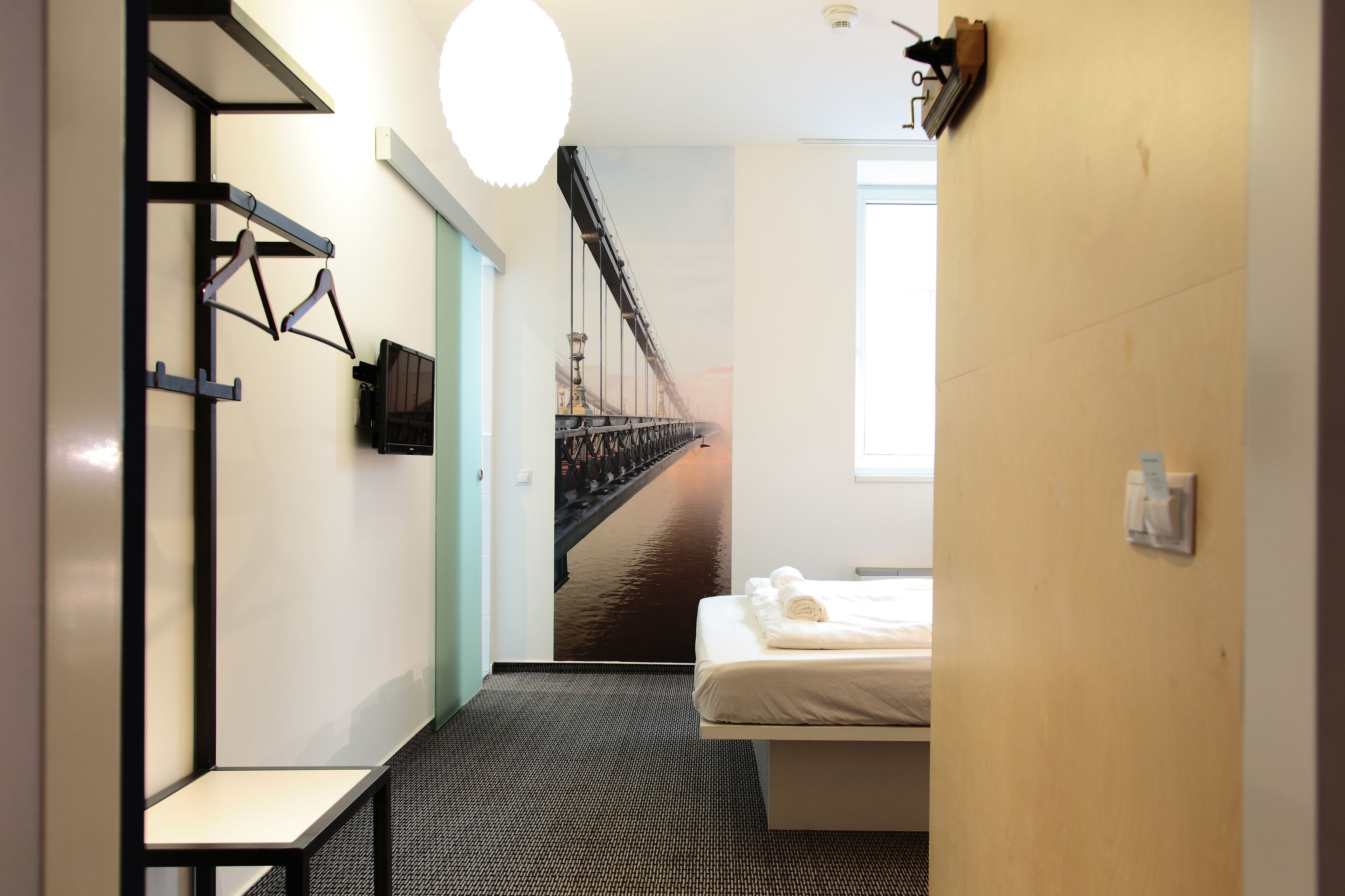 A hotel room with a bed, a desk, a hanging coat rack, and a large mural of a bridge.