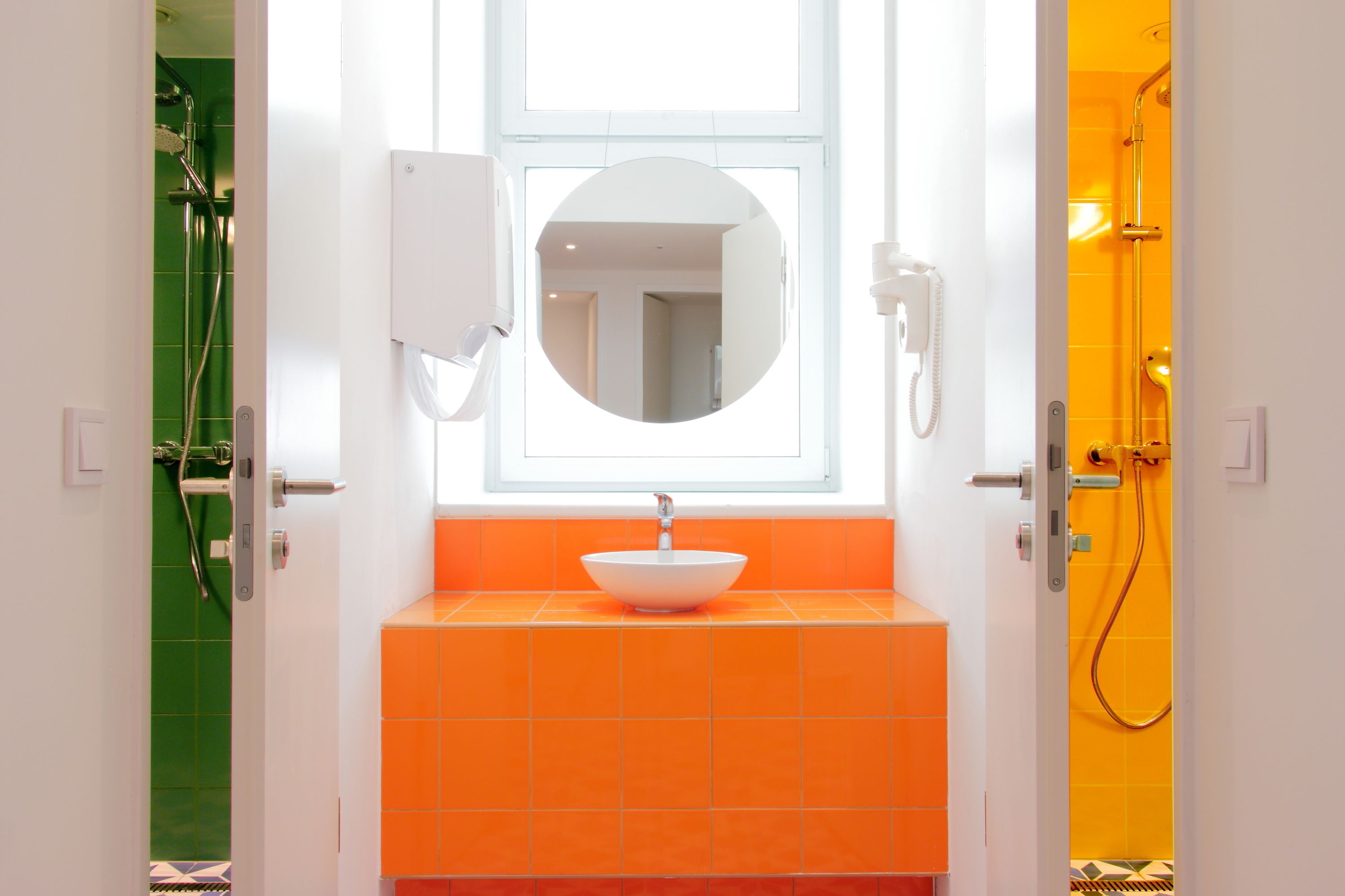 Un baño con un lavamanos blanco, azulejos naranjas, un espejo redondo y una ducha con puerta amarilla.