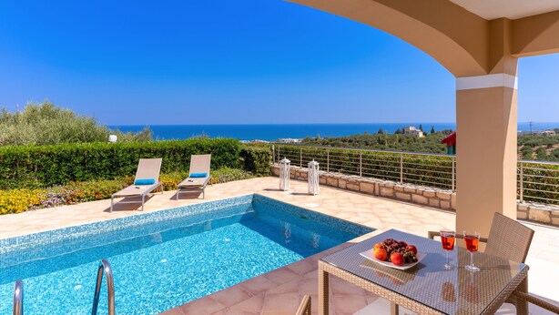 Zona de piscina con sillones de descanso y una mesa con bebidas y fruta, con vistas a un paisaje pintoresco.