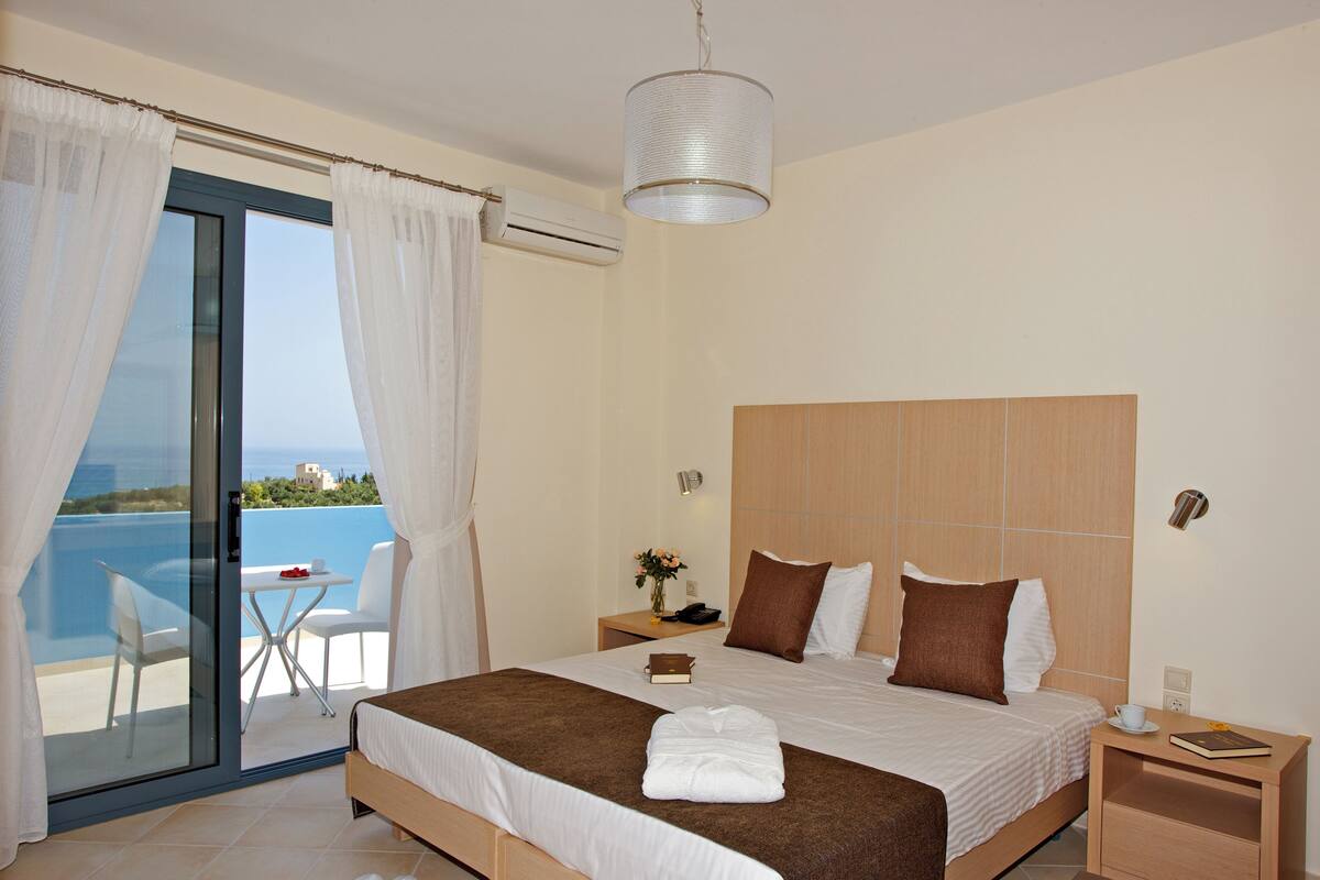 A hotel room with a bed, a nightstand, a bedside table, a lamp, a window with curtains, and a view of the sea.