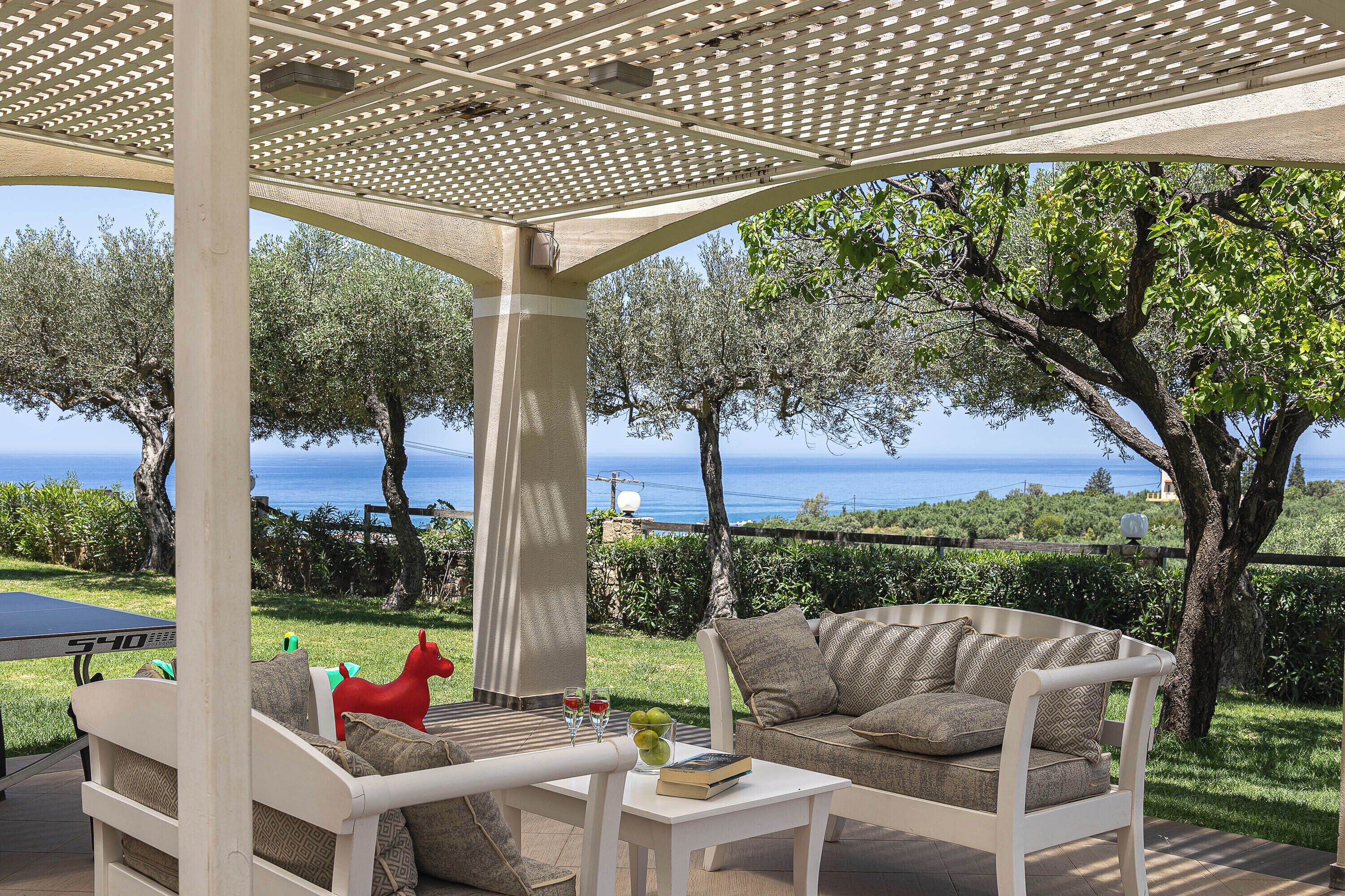 A covered outdoor seating area with a view of the sea, a table with a book and glasses, and a red horse decoration.