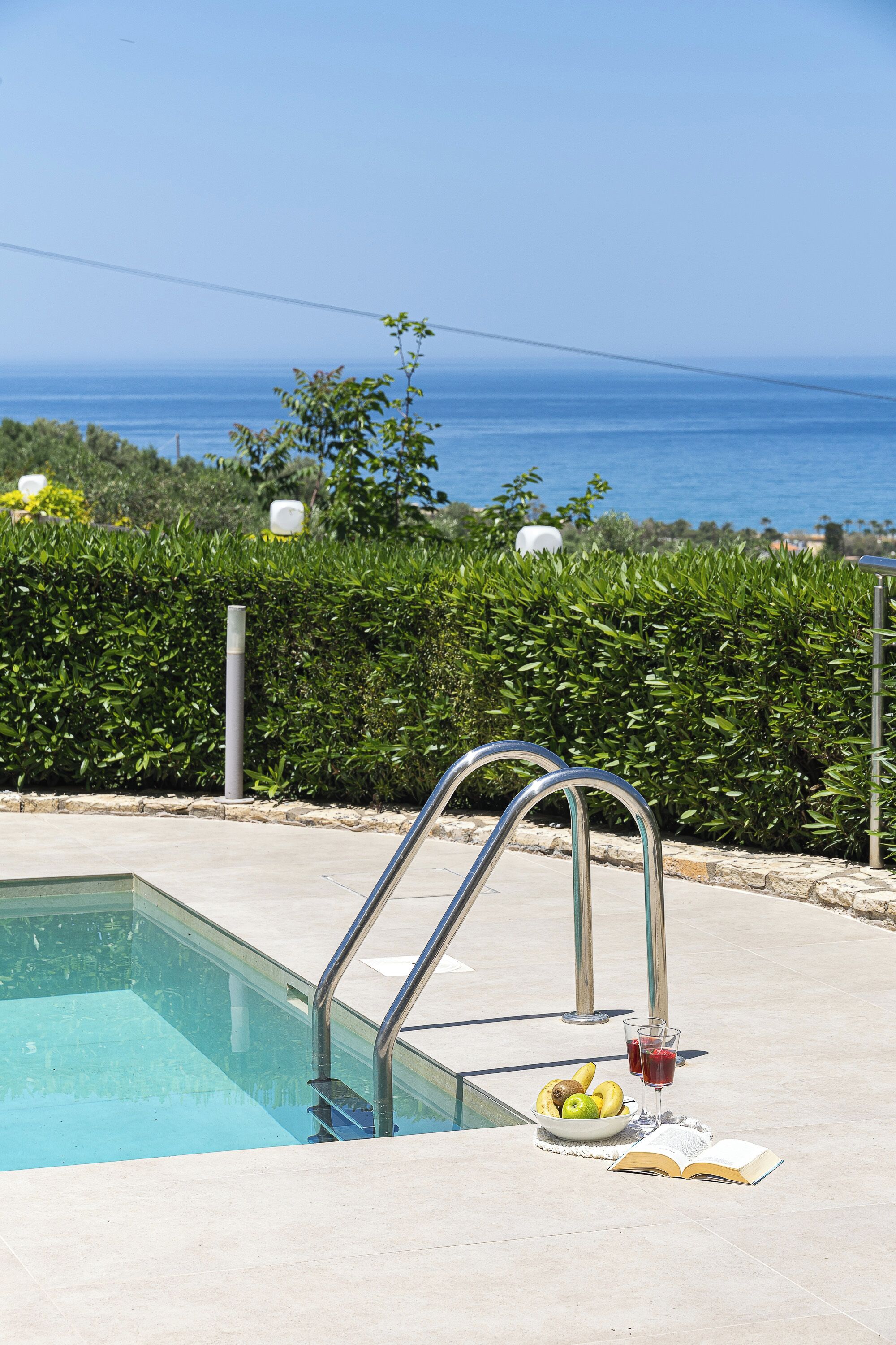A swimming pool with a metal ladder, a bowl of fruit, and a glass of red drink on the edge.
