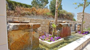 Una fuente de piedra con agua en cascada, rodeada de macizos de flores y un muro de piedra.