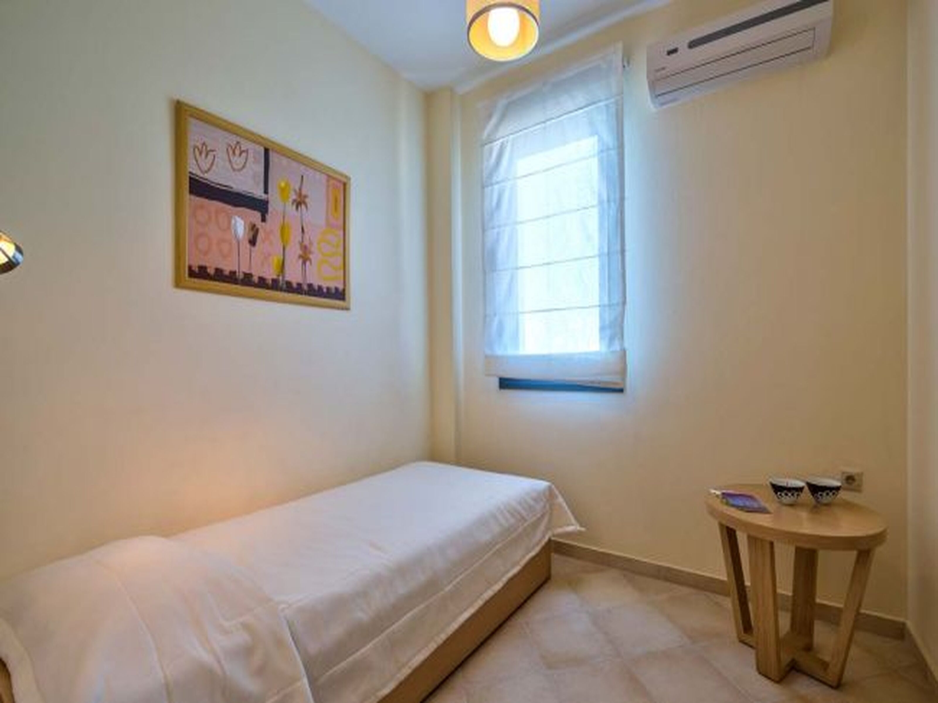 Habitació petita amb un llit individual, una finestra, una taula de nit i aire condicionat fixat a la paret.